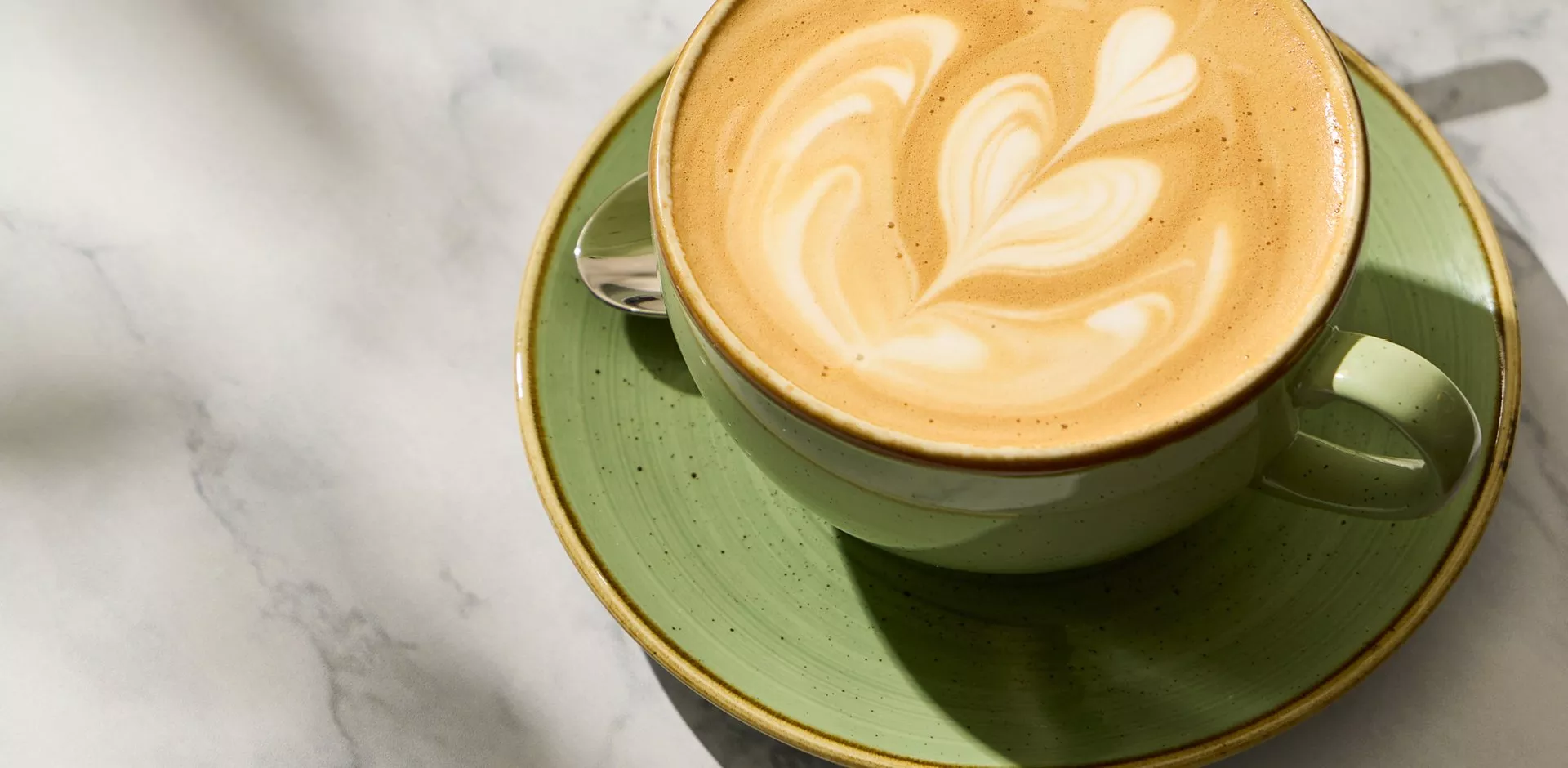 Latte with leaf latte art in a green ceramic cup and saucer, sunlit on a marble café table background
