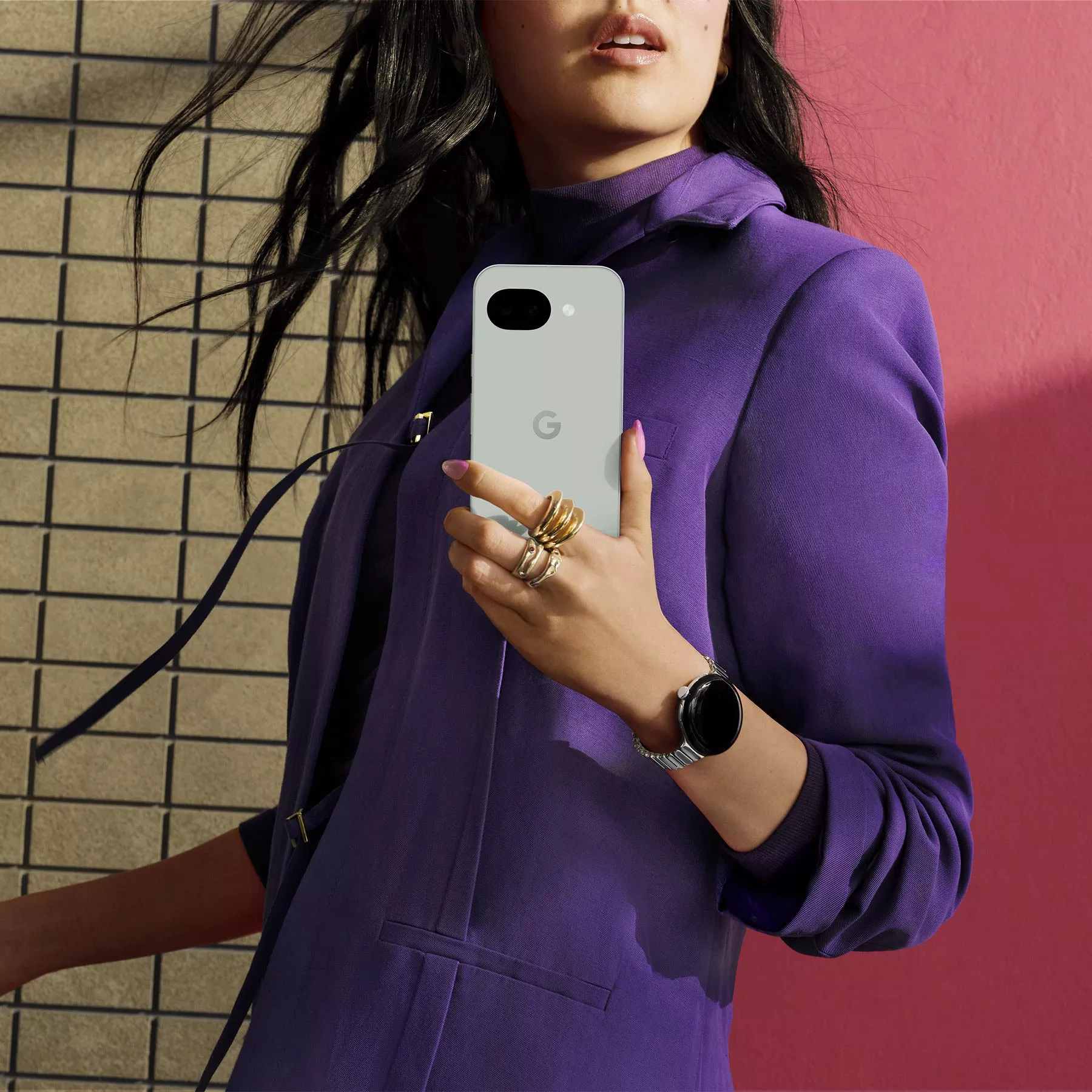 Woman in a purple outfit holding a smartphone with a prominent "G" logo, set against a pink and brick background.
