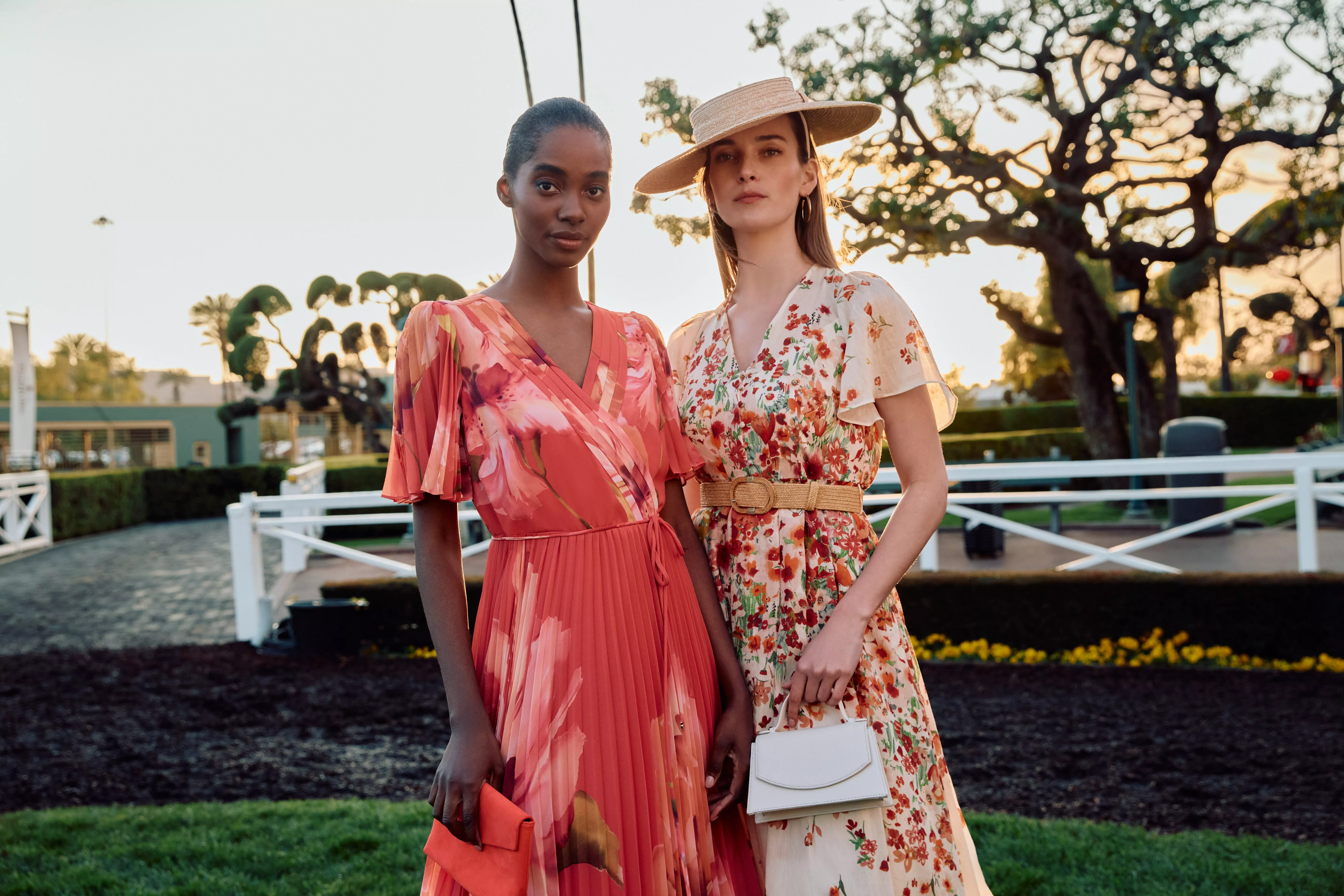 Two women in floral summer dresses and sun hat posing outdoors at sunset, holding clutch handbags at a garden venue