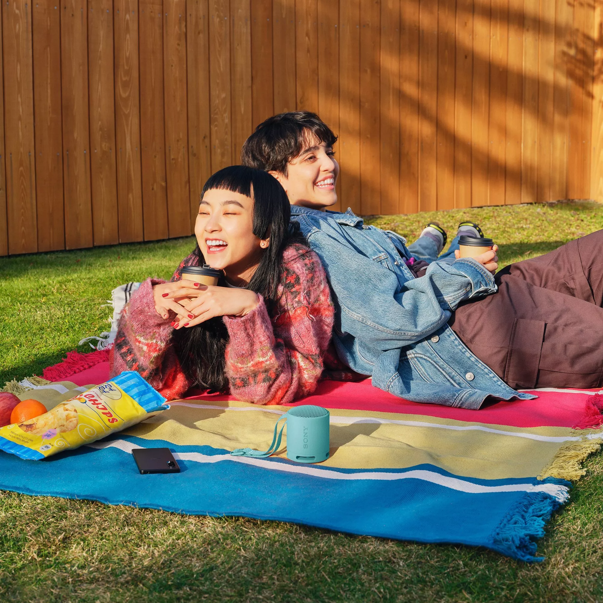 Two friends relaxing on a picnic blanket in the garden, holding takeaway coffees with snacks and a speaker nearby