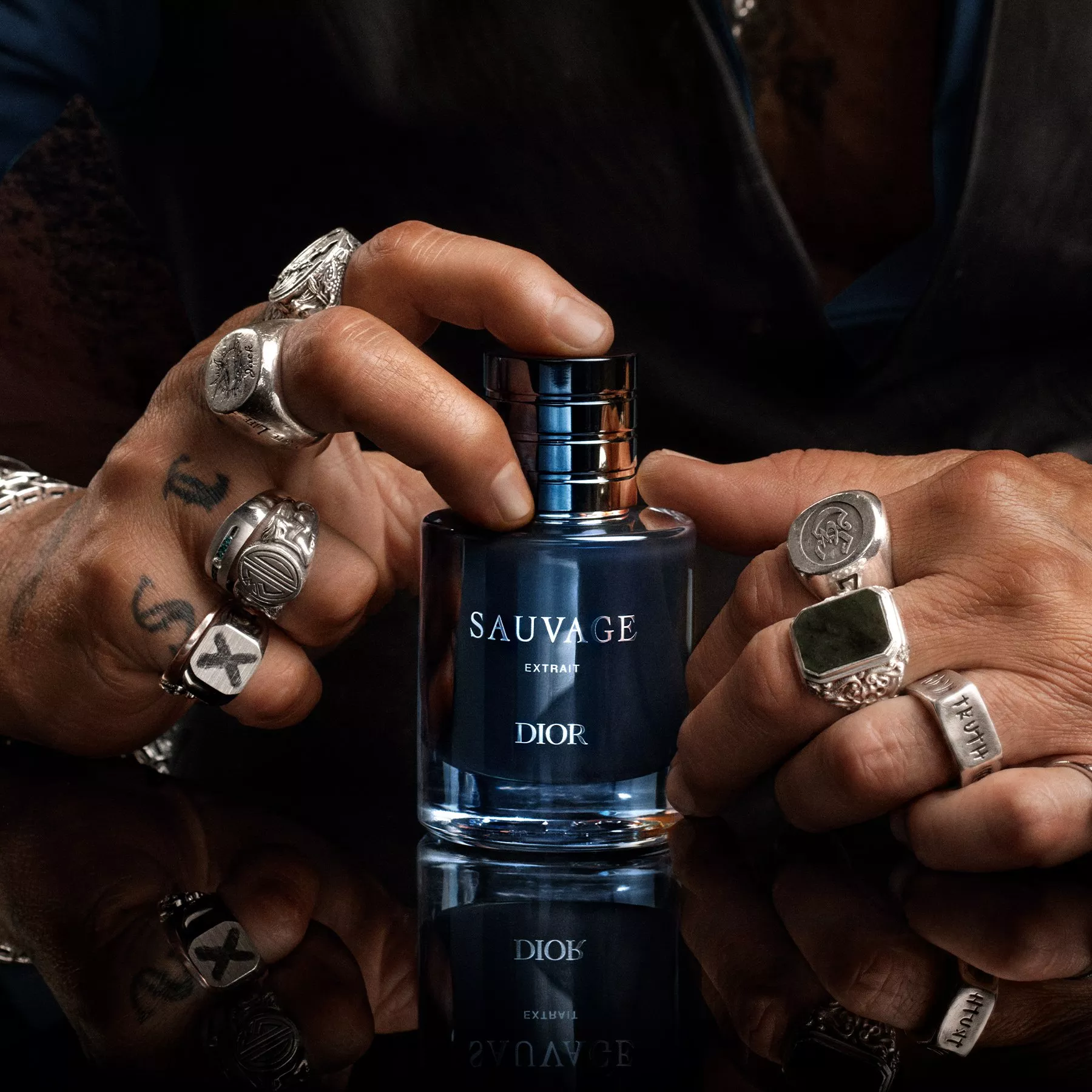 Close-up of hands with silver rings holding Dior Sauvage Extrait de Parfum bottle on reflective surface
