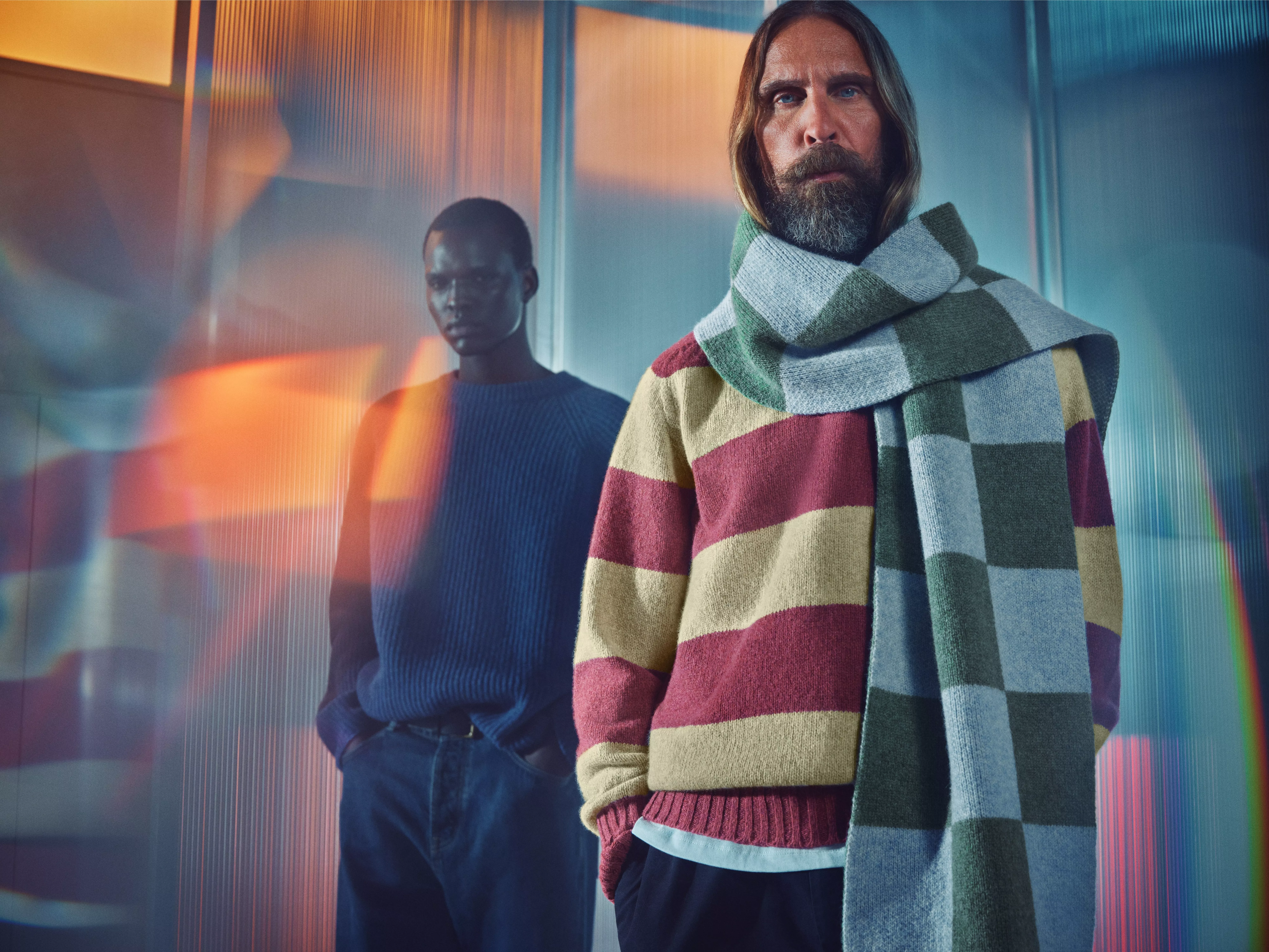Two men modeling winter apparel against a modern, iridescent backdrop. The man in the foreground wears a red and tan striped sweater and a large green and grey checkered scarf.