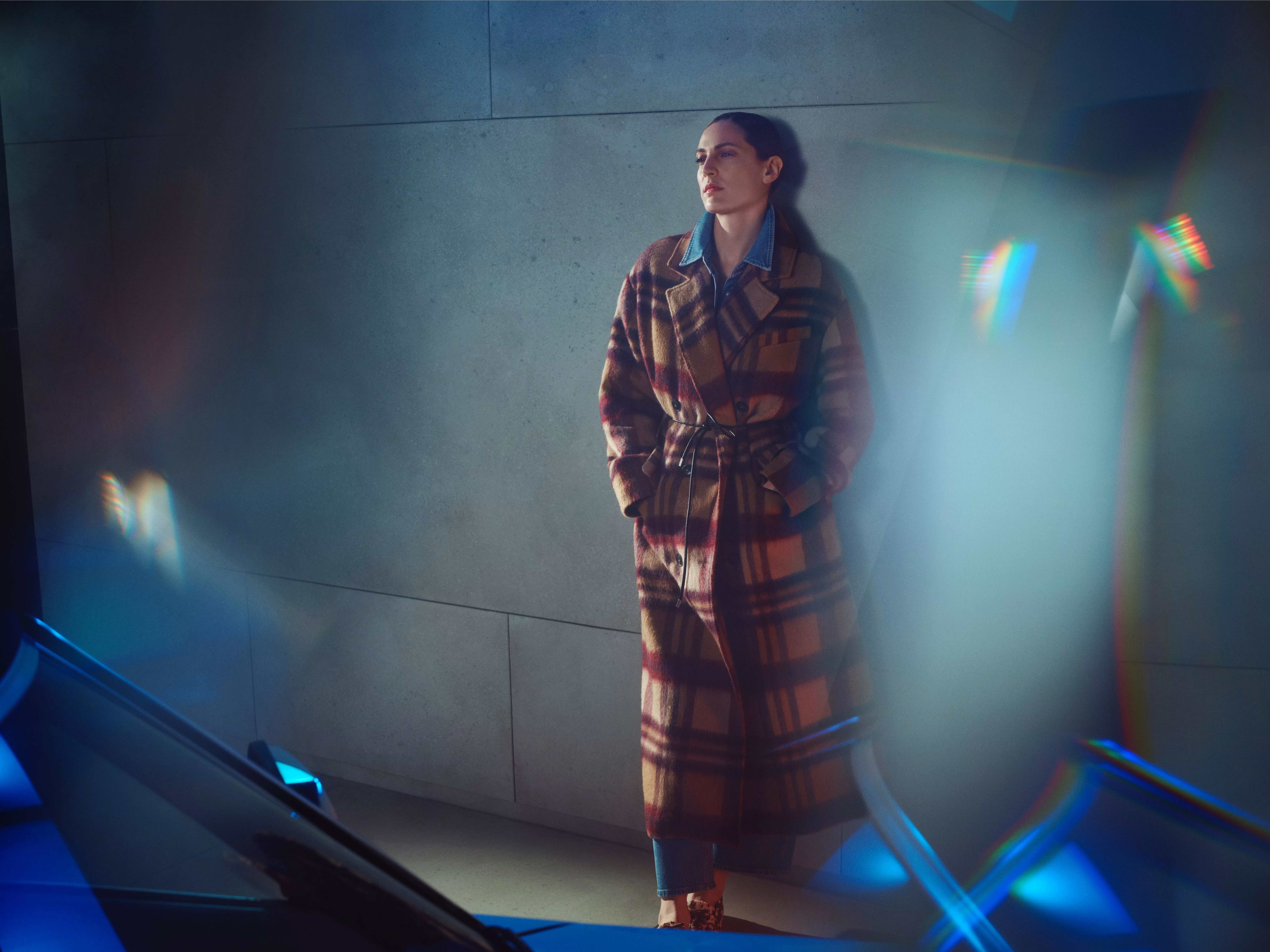 A woman wearing an earthy tone full length tartan mac coat against a grey tiled wall and blue lighting