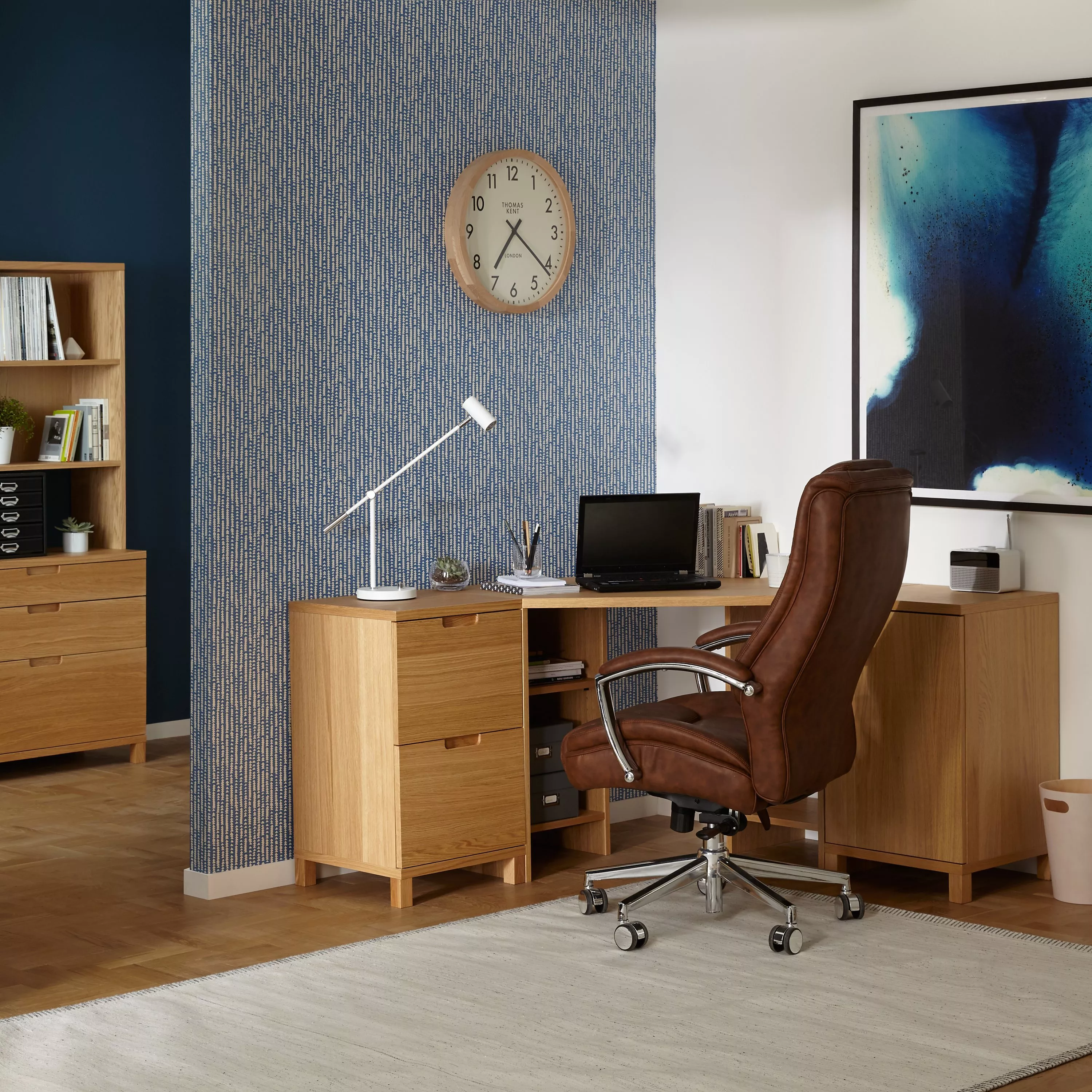 Modern home office with oak desk, leather swivel chair, blue patterned wallpaper and wall clock feature