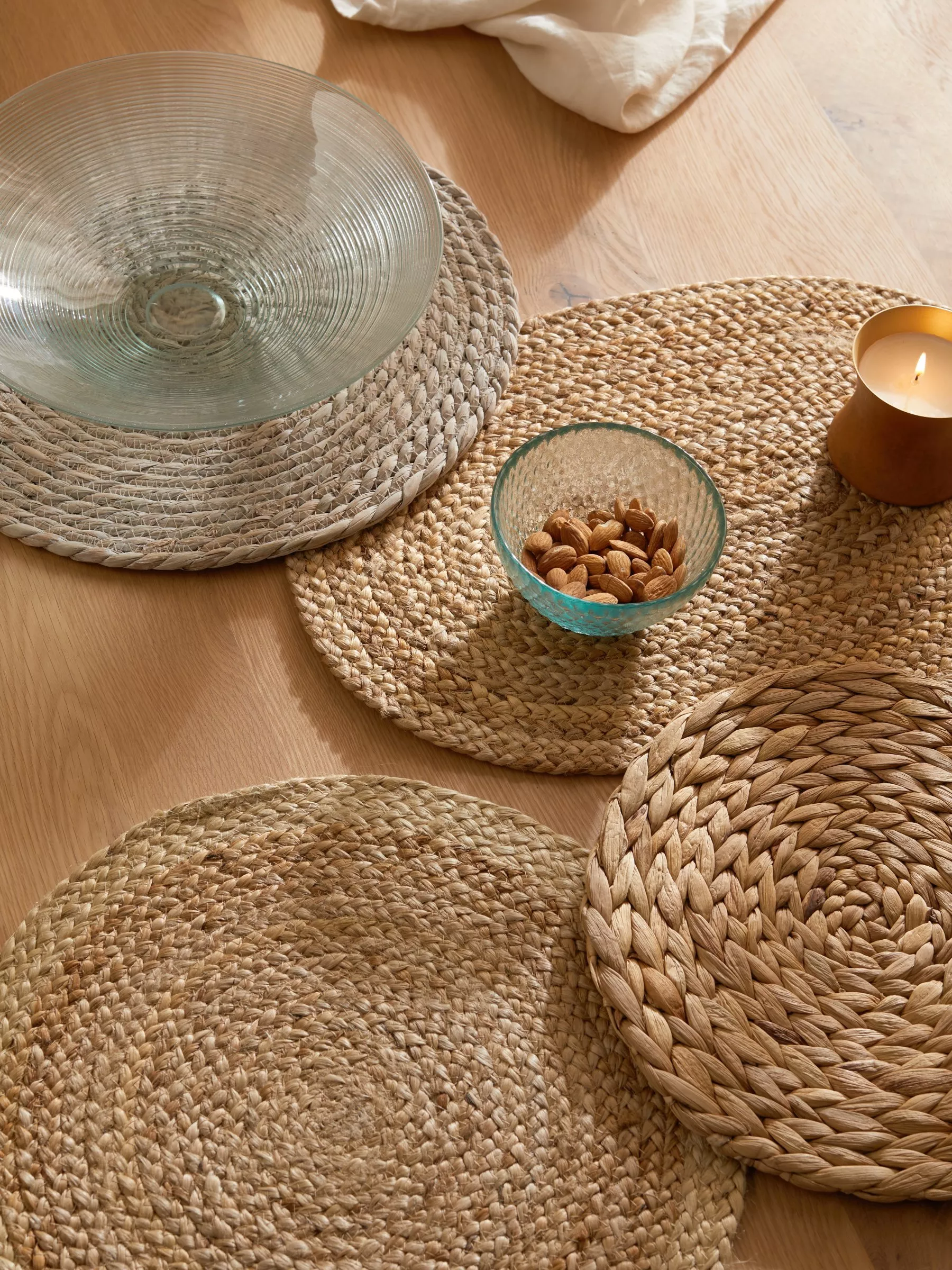 Natural woven jute placemats on wooden dining table with glass bowl, almonds in dish and lit candle decor