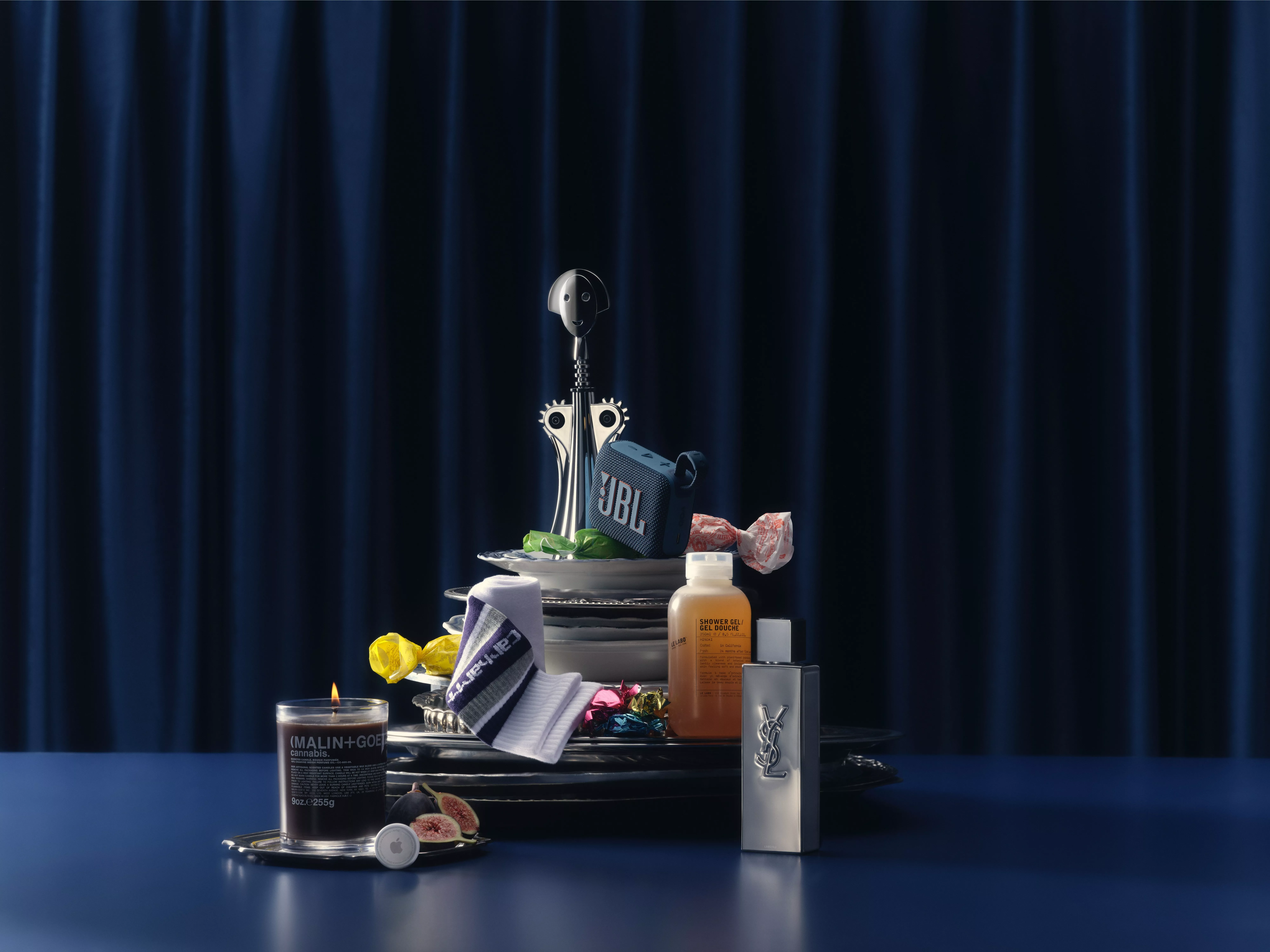 A stack of plates featuring various items, including a JBL speaker, a candle, a bottle of YSL perfume, and other gifts, set against a dark blue curtain backdrop