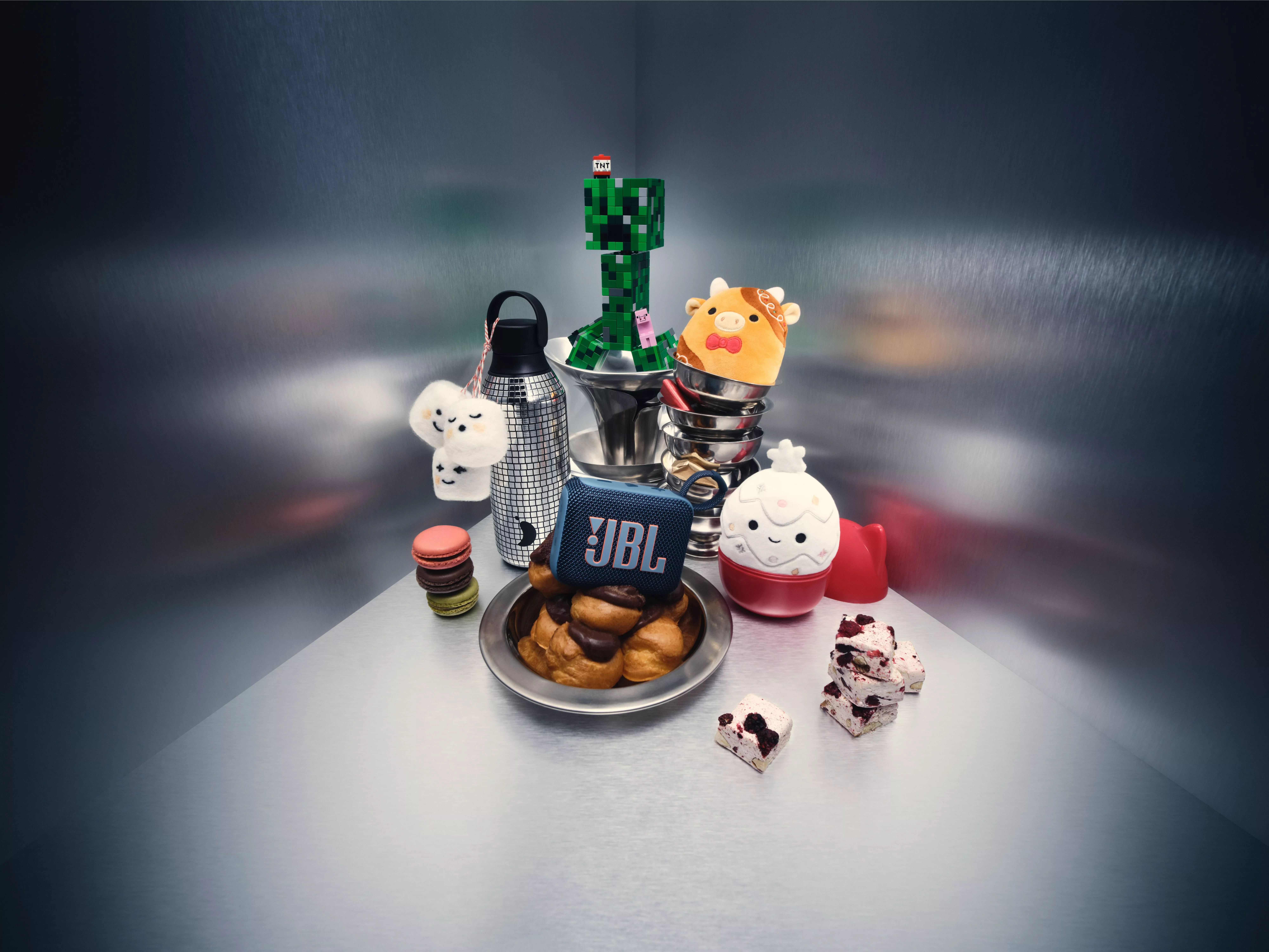 A product arrangement featuring a variety of items including a JBL speaker, a Minecraft Creeper figure, various plush toys, and a plate of pastries, all on a metallic background