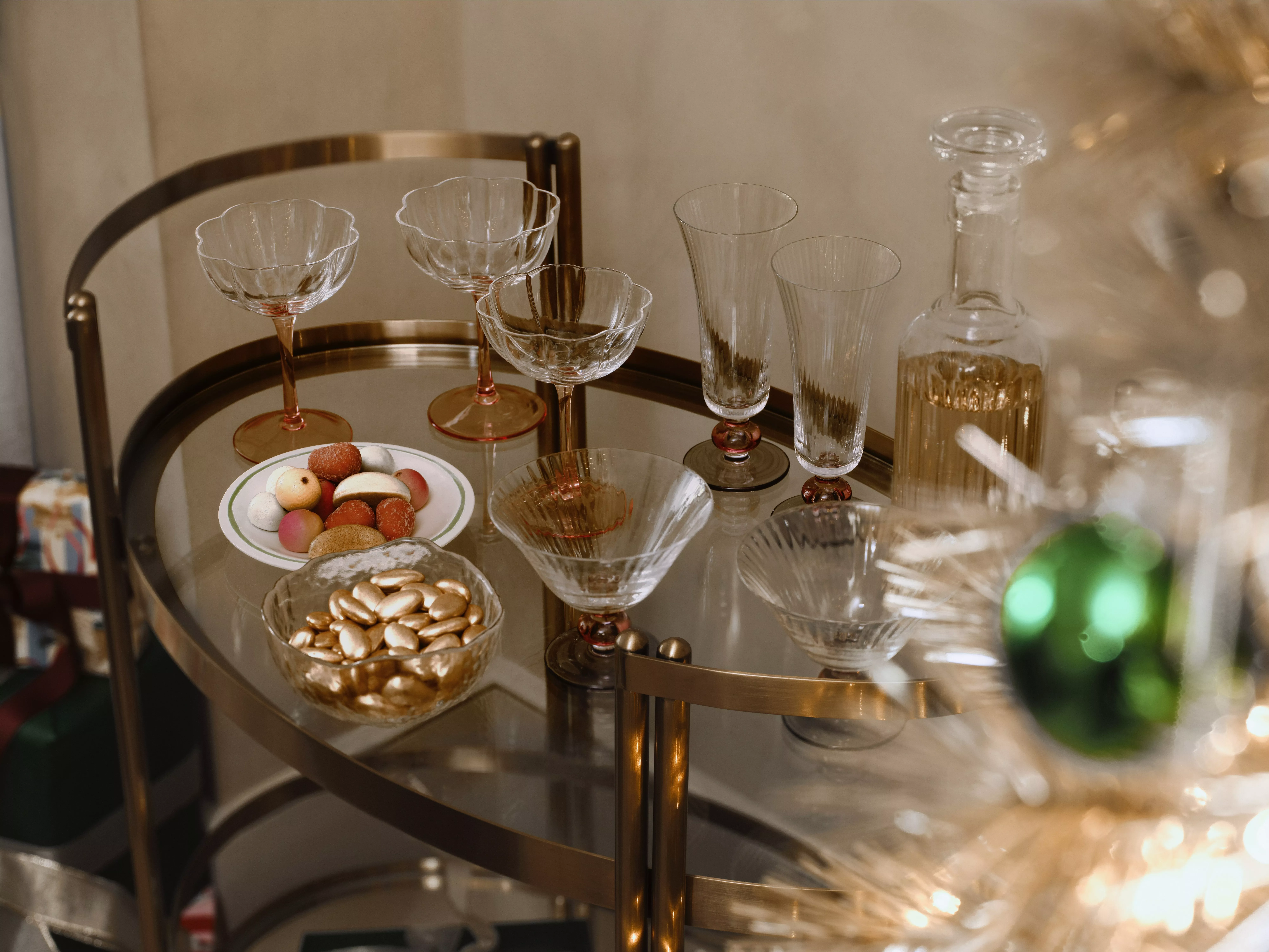 a gold-framed glass bar cart holds elegant coupes, flutes, a decanter, and bowls of candy and fruit next to a decorated tree.