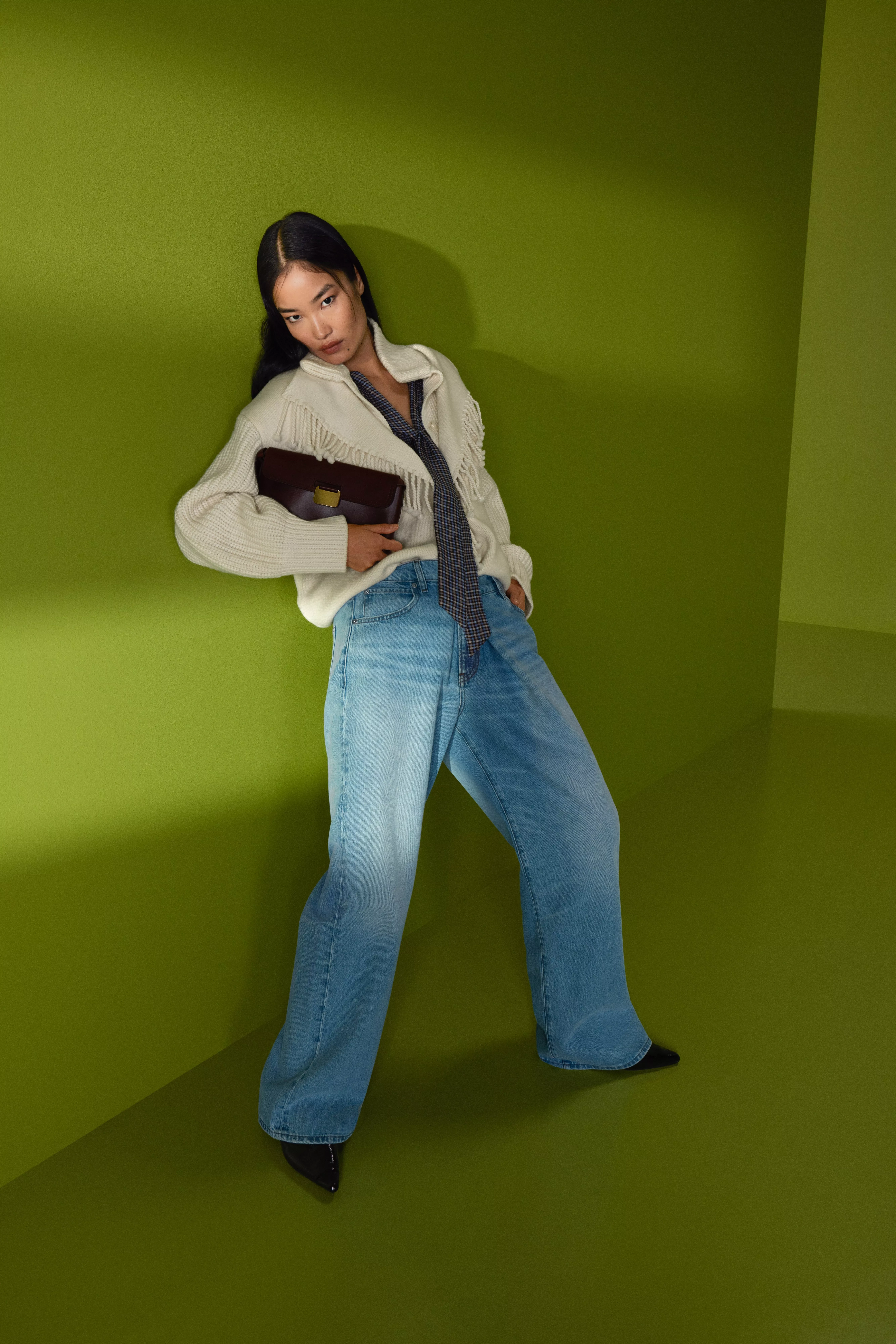 Fashion model in cream fringed cardigan and wide-leg jeans, holding brown clutch against green backdrop