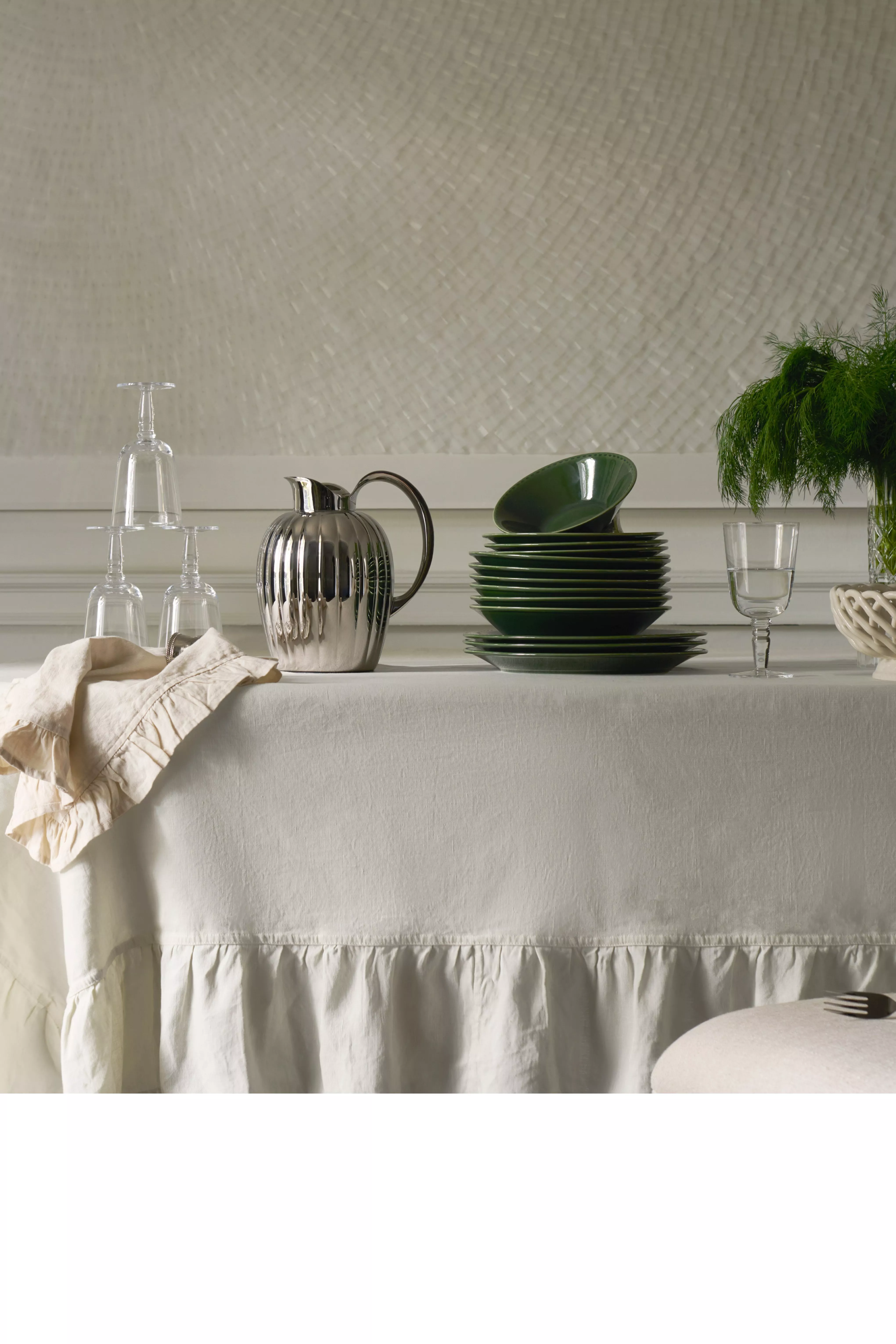 Elegant dining table setting with green crockery, silver jug, glassware and vase of greenery on linen cloth