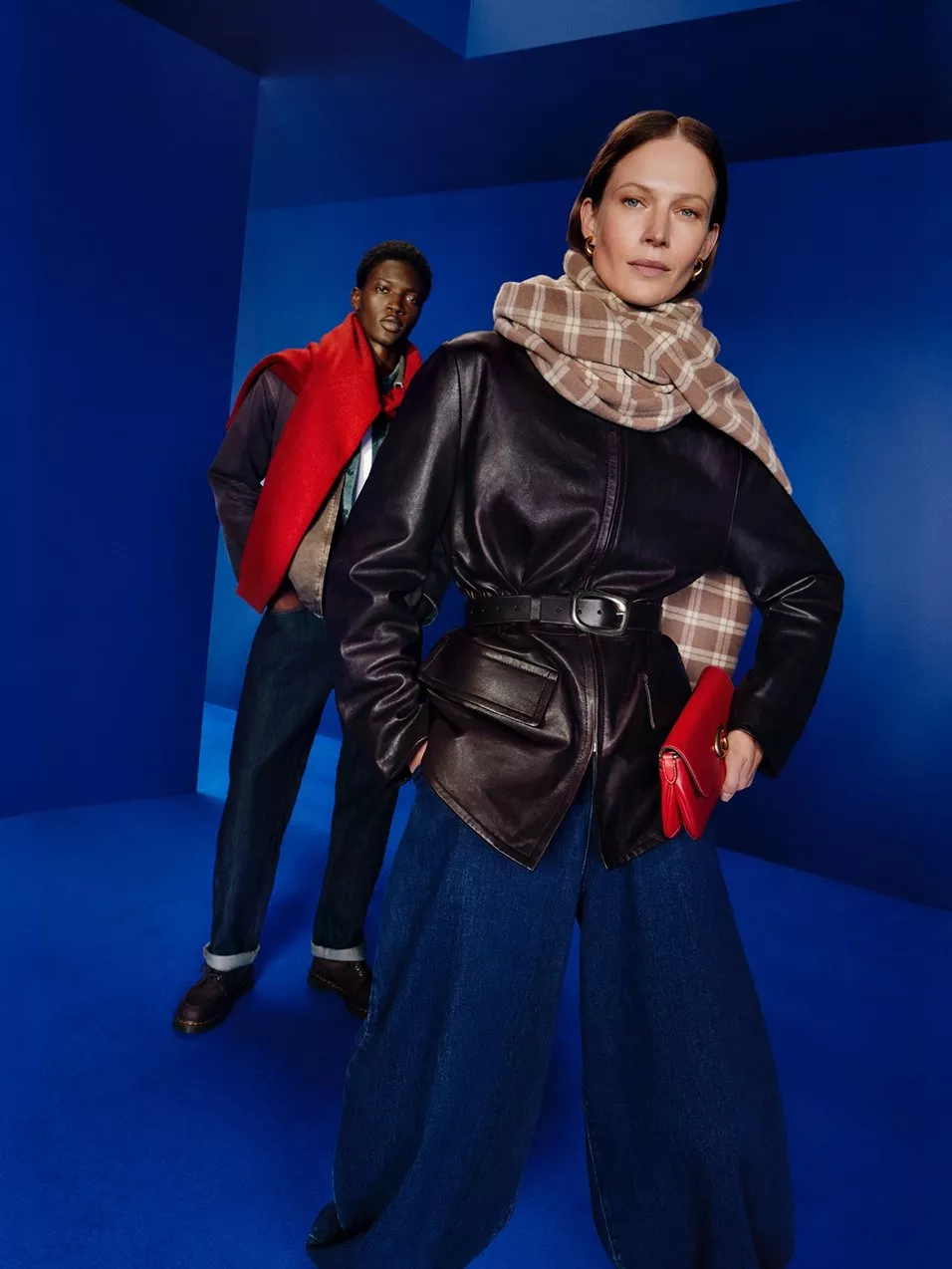 Fashion models in leather jacket and plaid scarf, with red accessories, posing in a blue studio backdrop