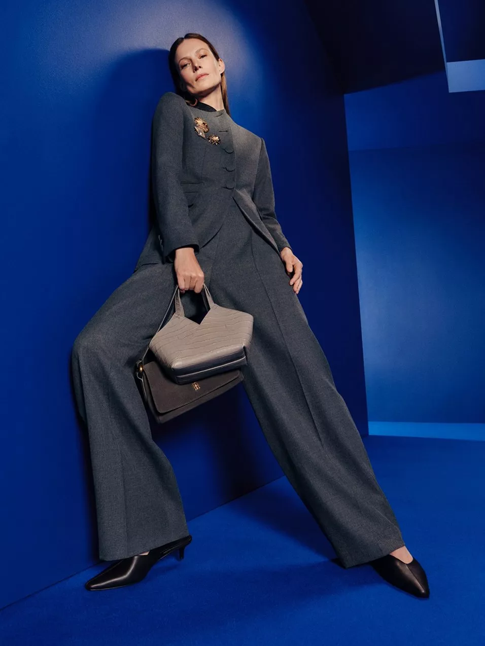 Fashion model in grey tailored jumpsuit holding designer handbags, posing against bold blue studio backdrop