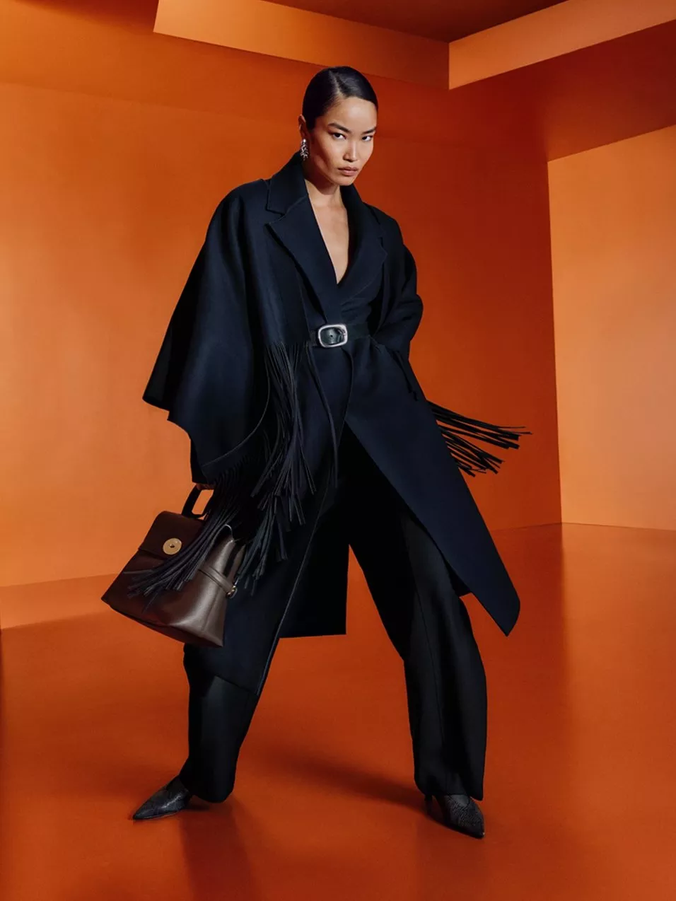 Fashion model in black belted fringed coat and trousers, holding brown handbag against orange studio backdrop