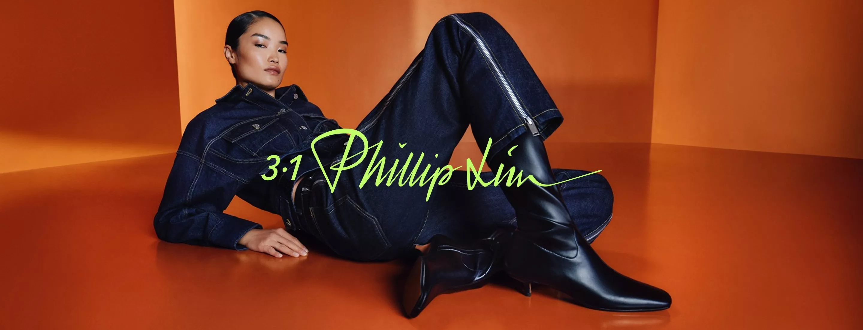Fashion model in dark denim jacket and jeans with black ankle boots, reclining on orange studio set, 3.1 Phillip Lim logo