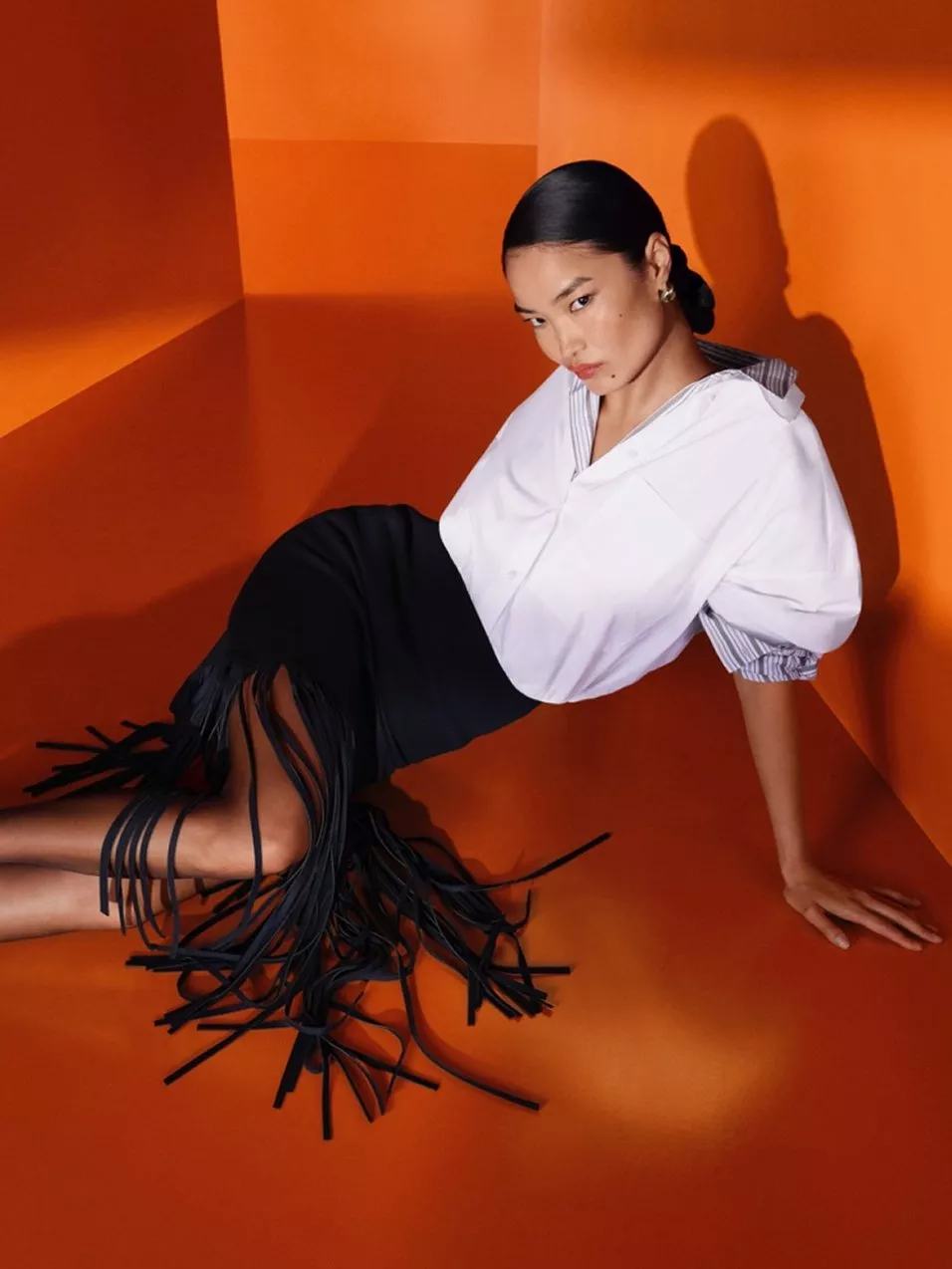 Fashion editorial model in white blouse and black fringed skirt, posing in vibrant orange studio set