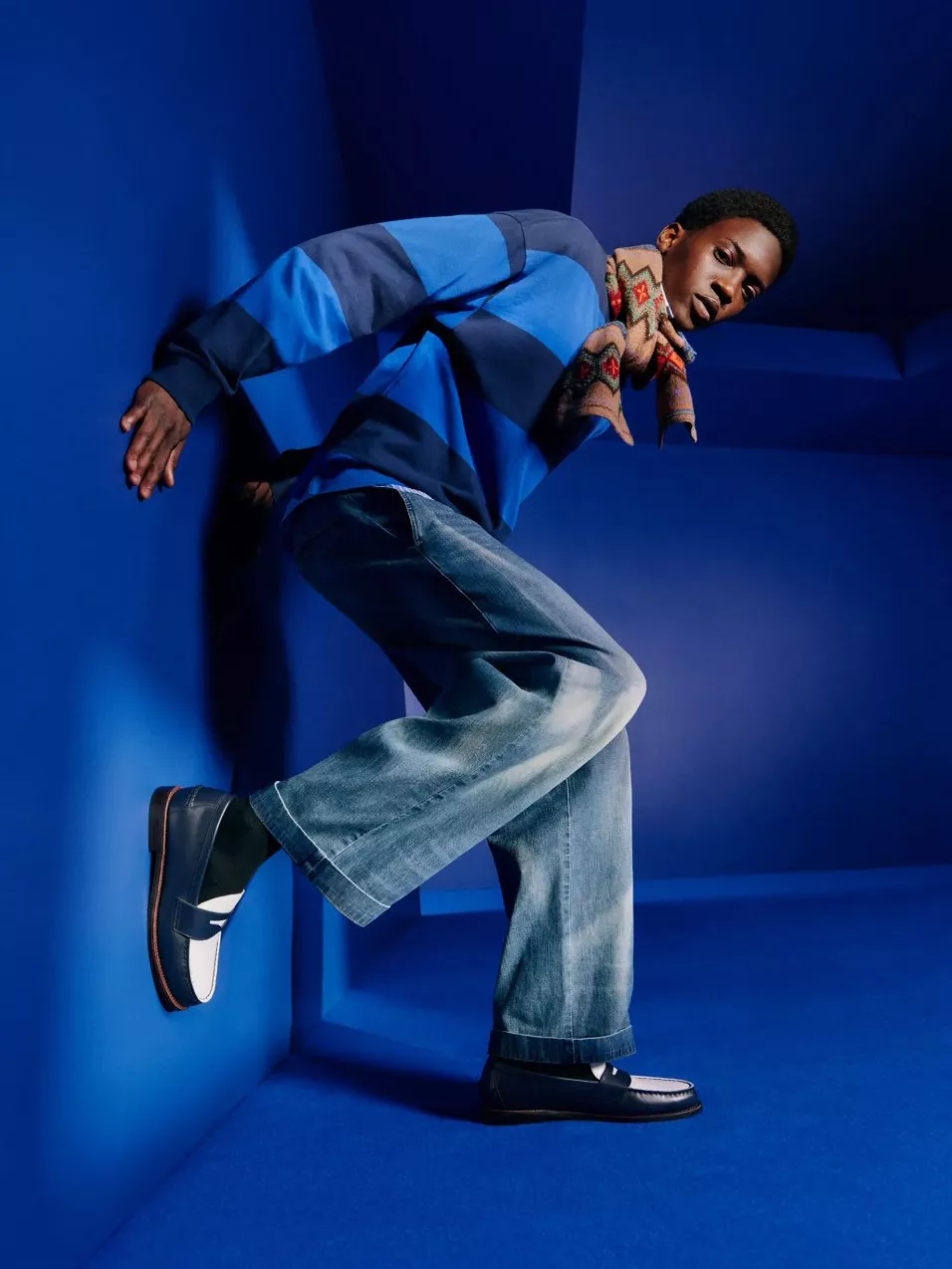 Fashion portrait of man in blue striped jumper, wide-leg jeans and loafers, posing in a blue studio set