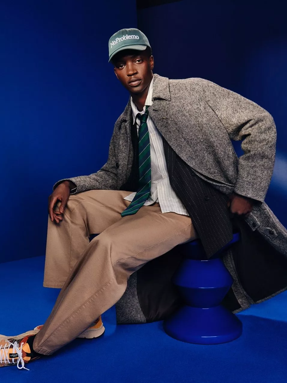 Men’s smart casual fashion portrait: model in grey overcoat, striped tie and cap, seated on blue set