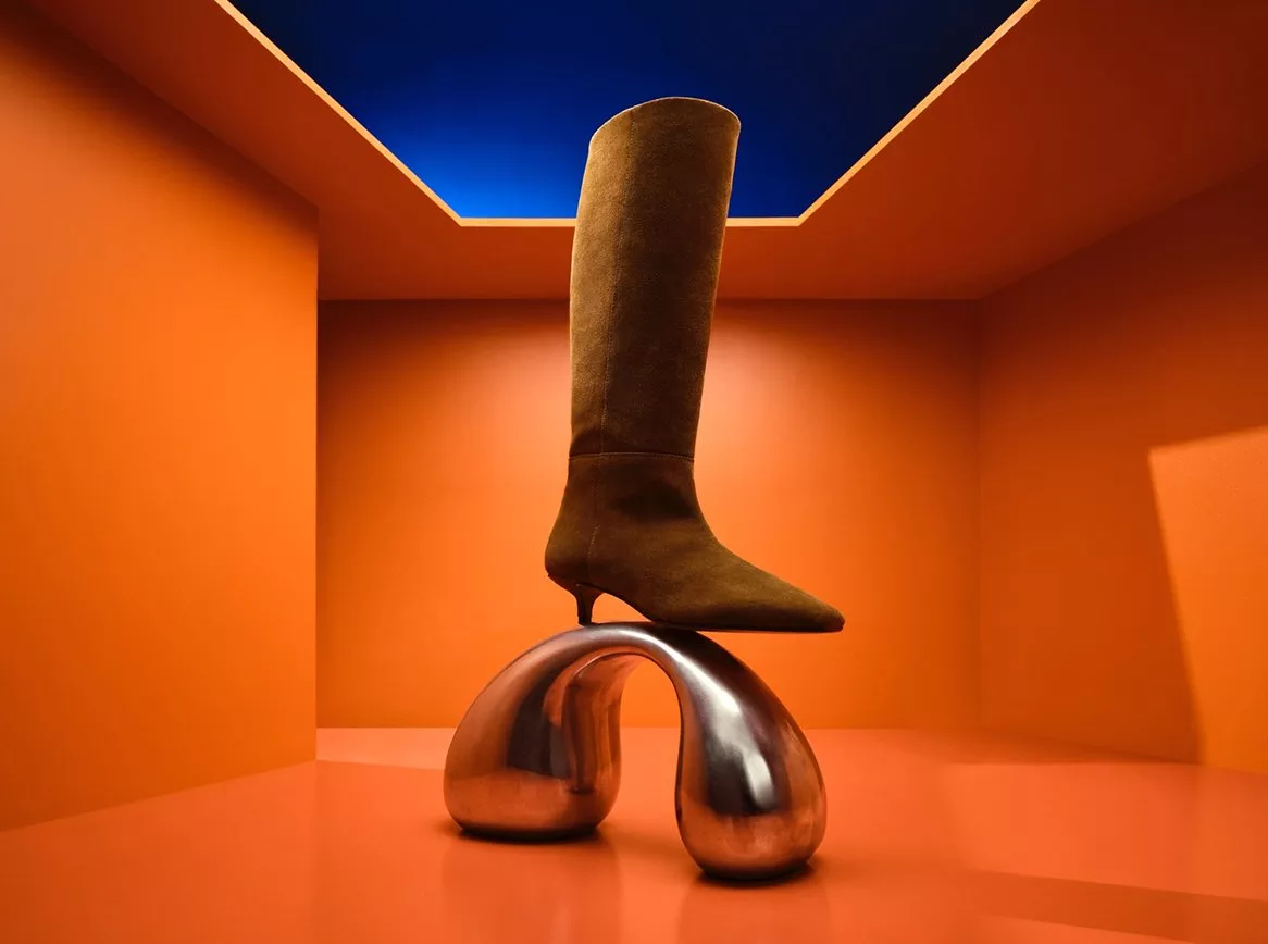 Brown suede knee-high heeled boot on polished chrome arch pedestal in a bold orange studio interior.