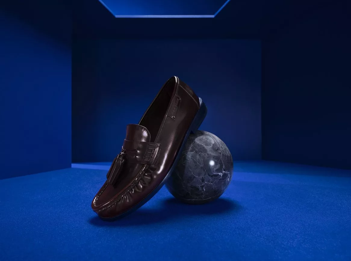 Dark brown leather tassel loafer propped on a marble sphere in a blue studio, luxury men’s footwear shot