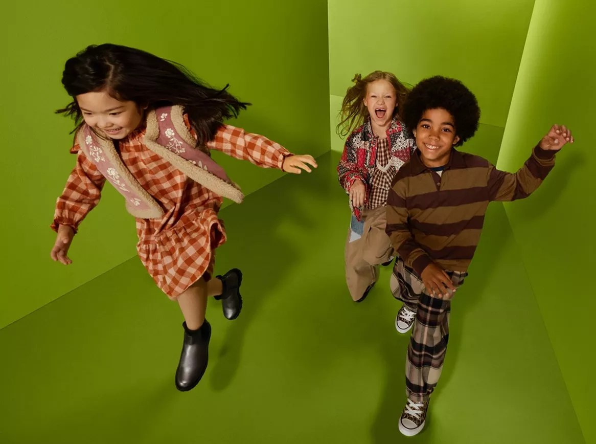 Three children running and smiling in a green studio, wearing autumn outfits in a playful fashion shoot