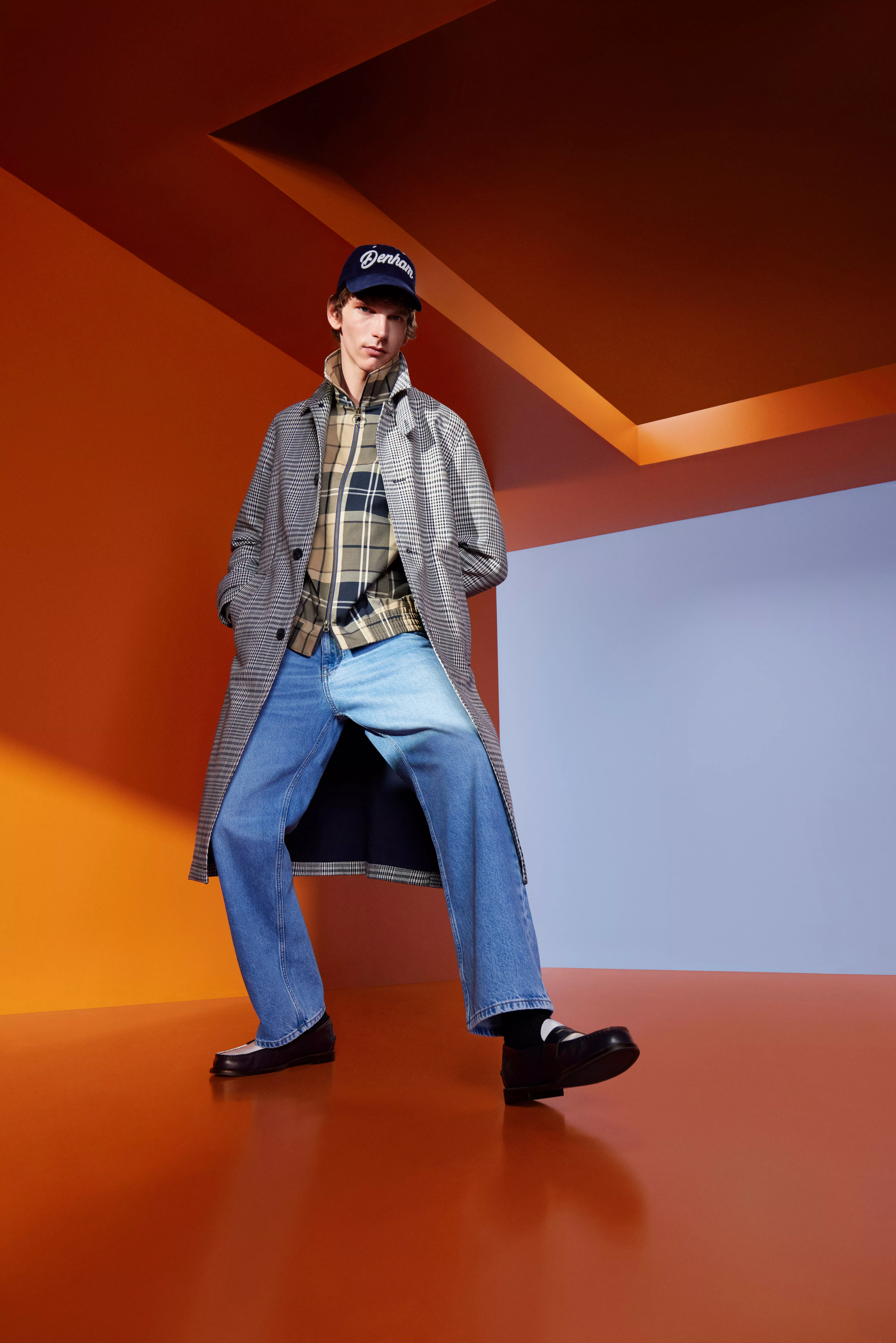 Male fashion model in checked overcoat, plaid shirt and wide-leg jeans, posing in bold orange studio set