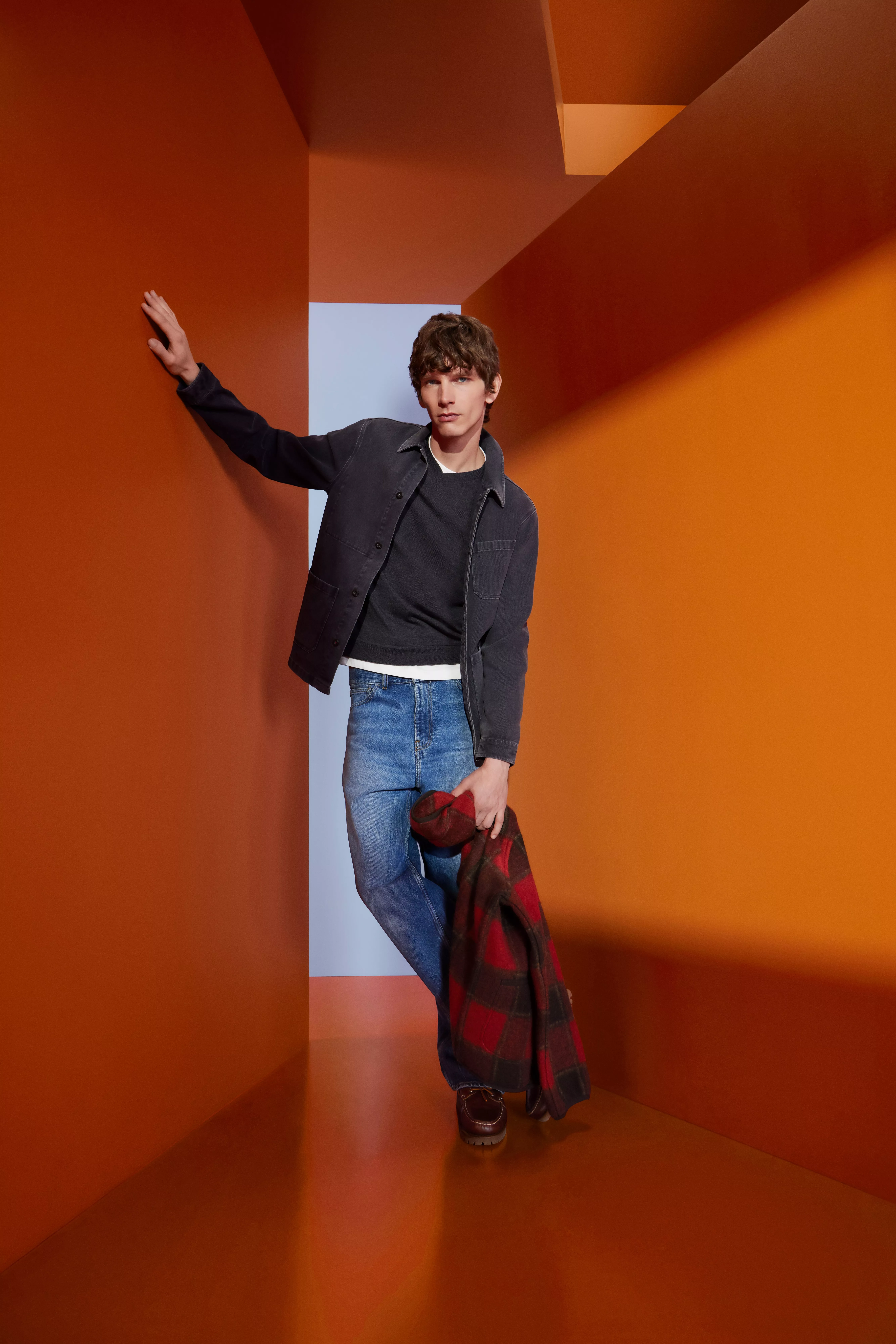 Male model in dark jacket and jeans holding tartan coat, posing in an orange geometric studio corridor