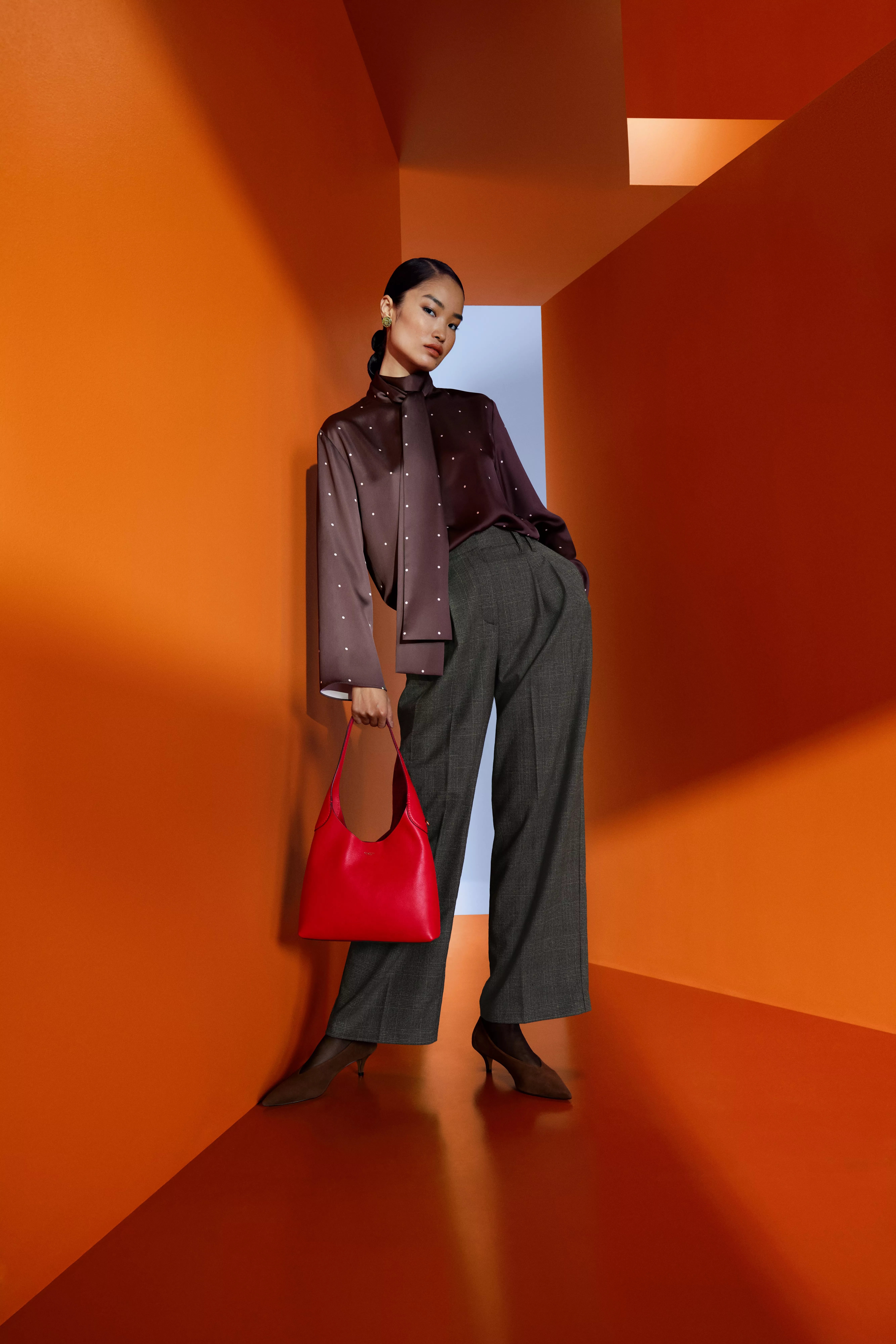Fashion model in brown blouse and wide-leg trousers holding red handbag, posing in vibrant orange studio corridor