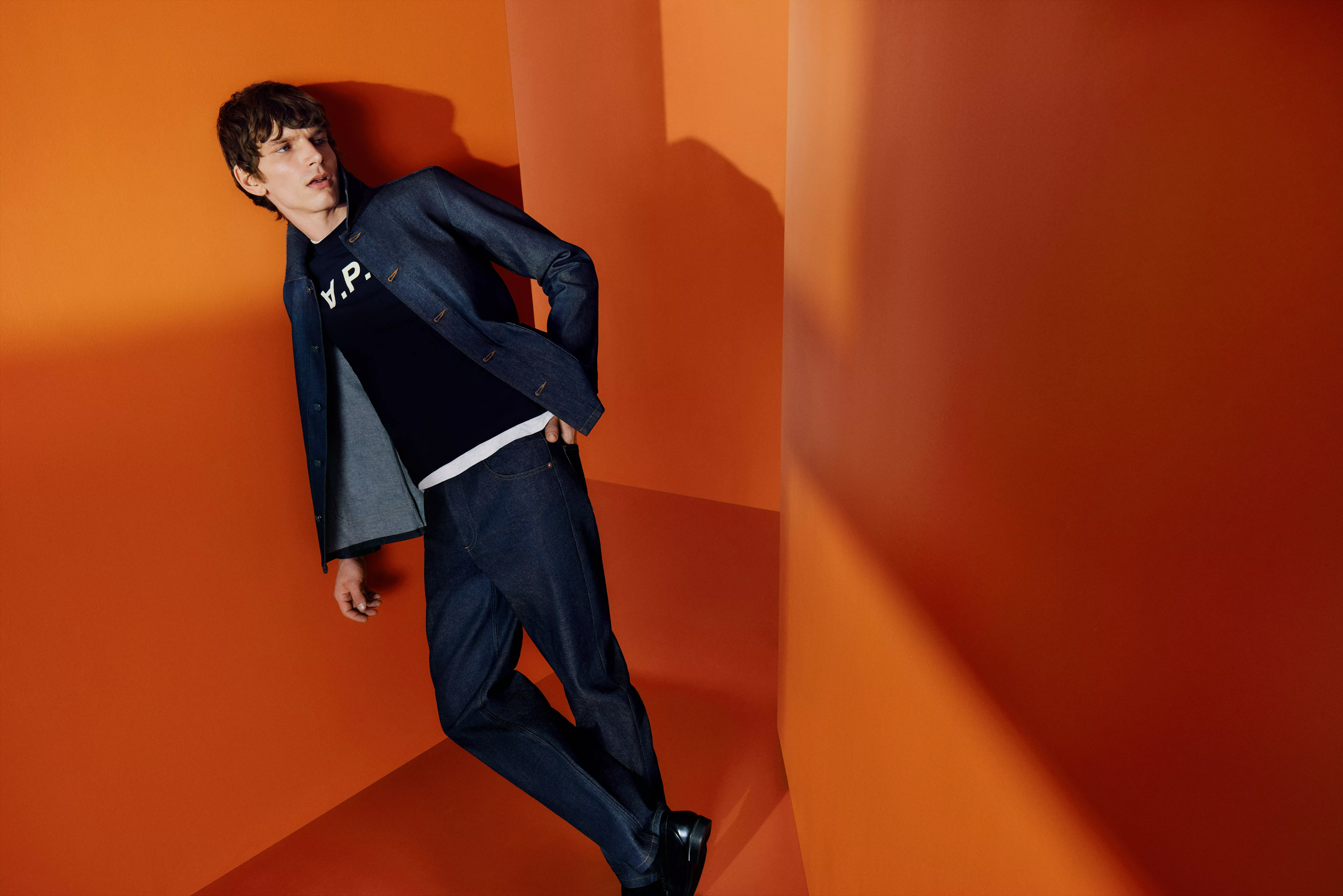 Fashion model in dark denim jacket and jeans, leaning against orange wall in modern studio photoshoot