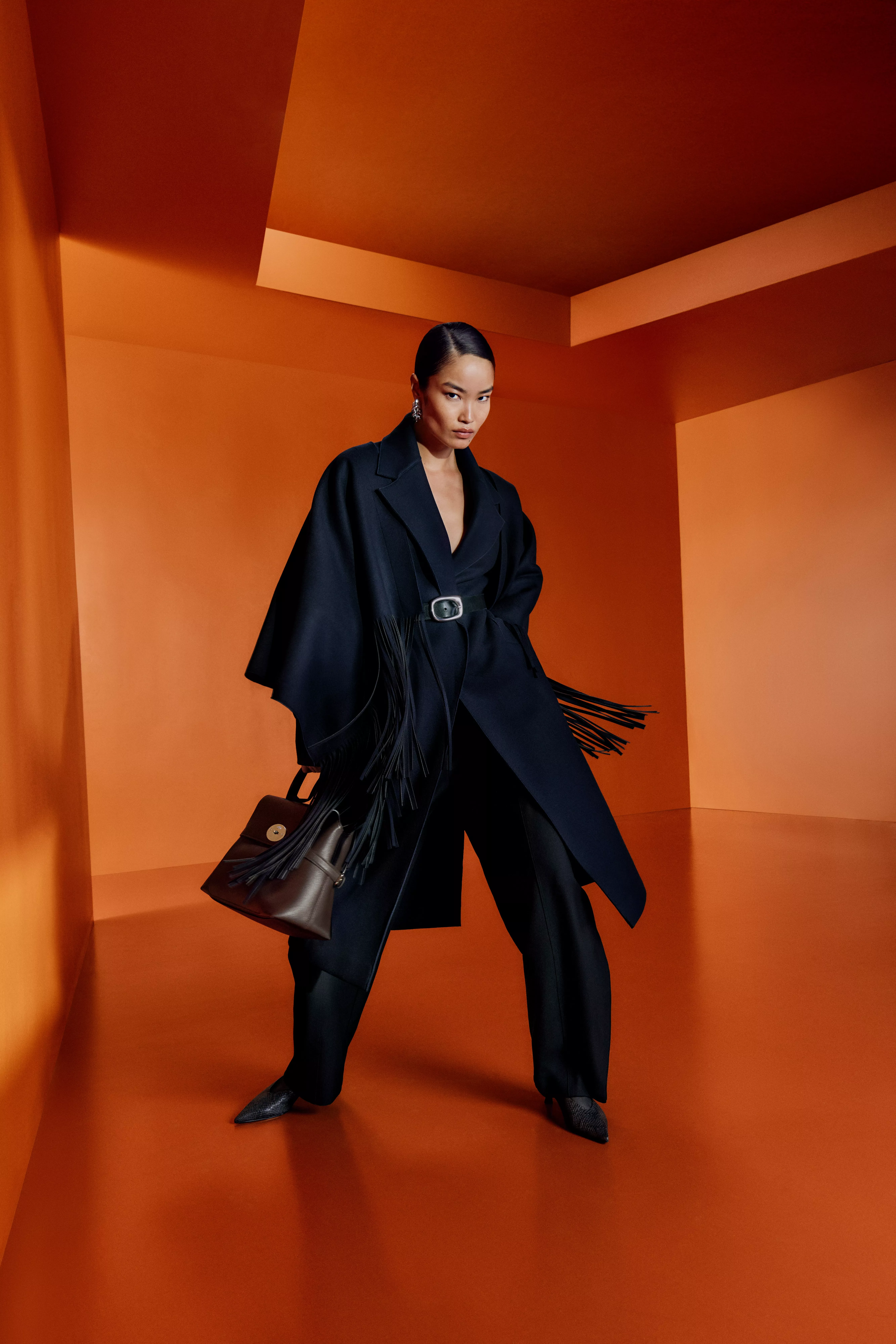 Fashion model in black belted coat with fringe, holding brown handbag, posing in orange studio set