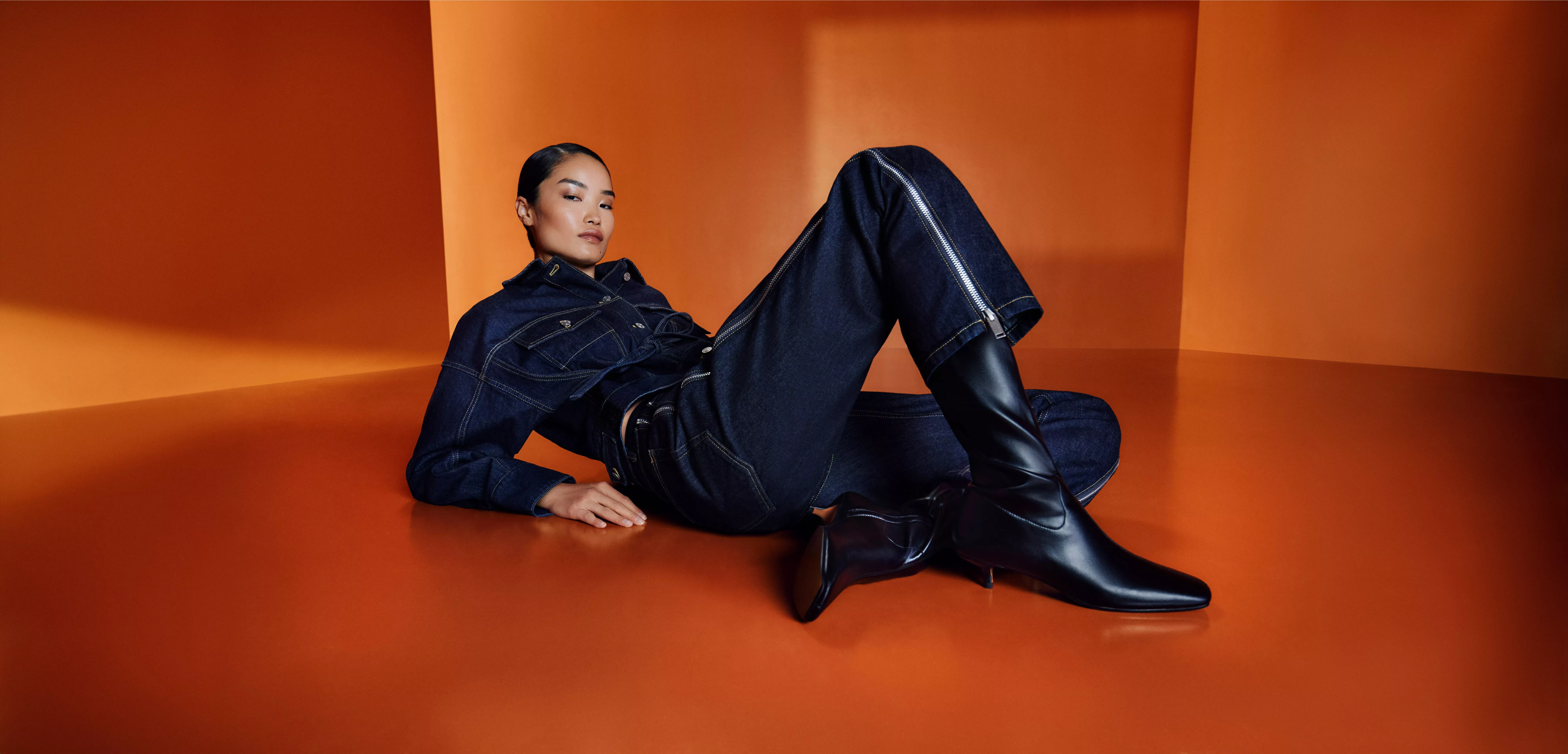 Fashion portrait of woman in black dress with white lace trim and knee-high boots, seated on modern chair.