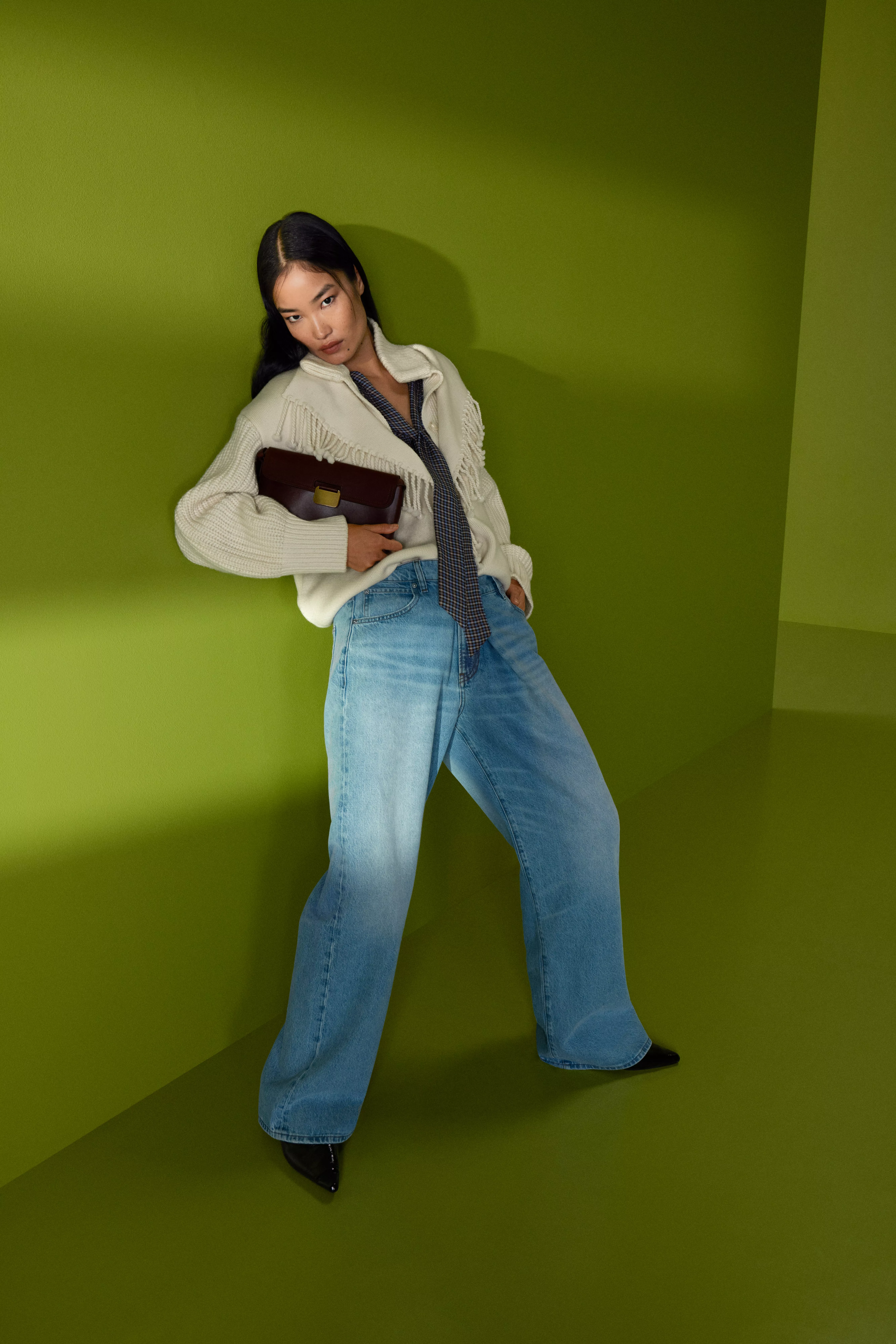 Fashion model in fringe cream cardigan and wide-leg jeans, holding brown clutch, posing on green backdrop