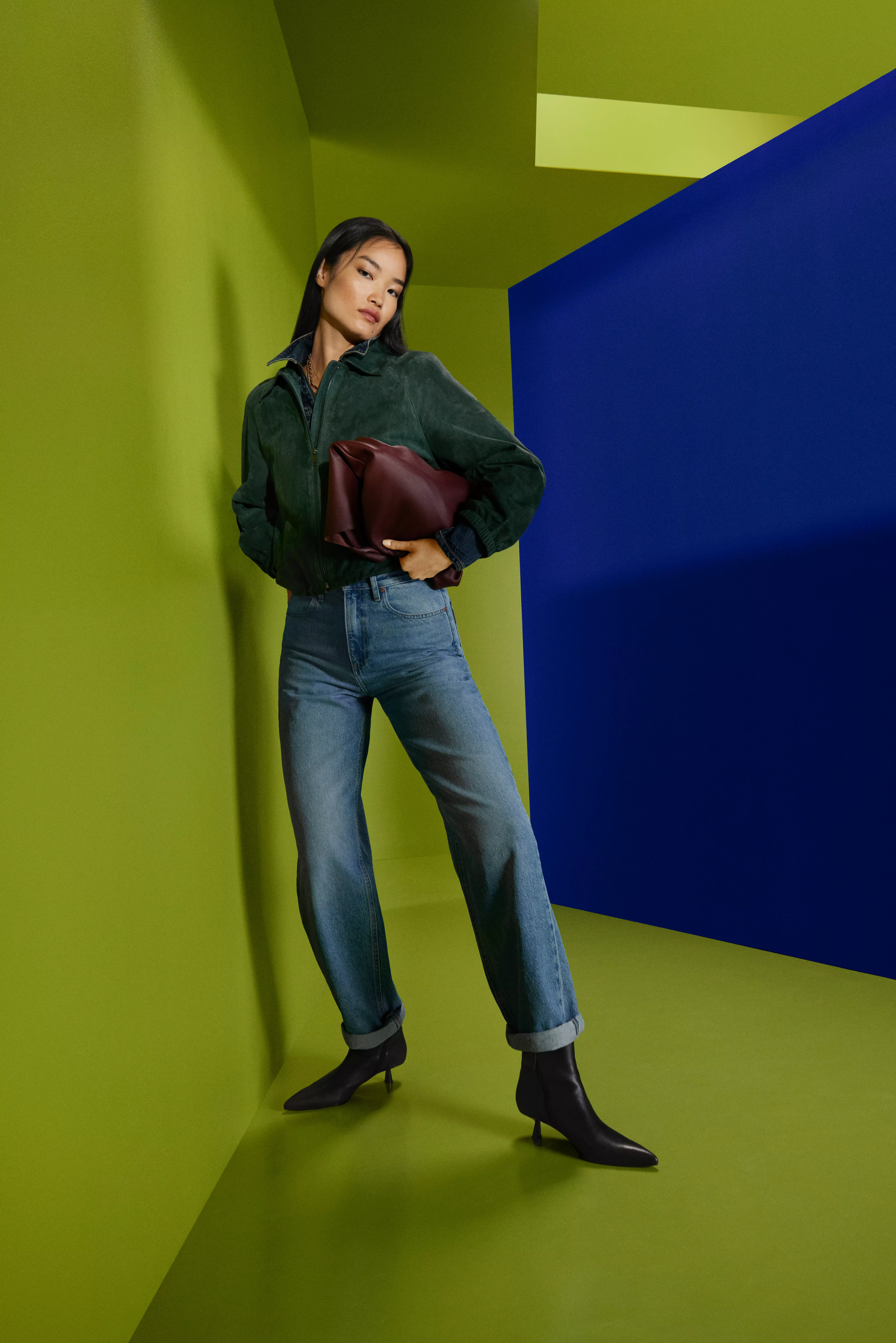 Fashion model in green jacket and blue jeans holding burgundy clutch in modern green and blue studio set