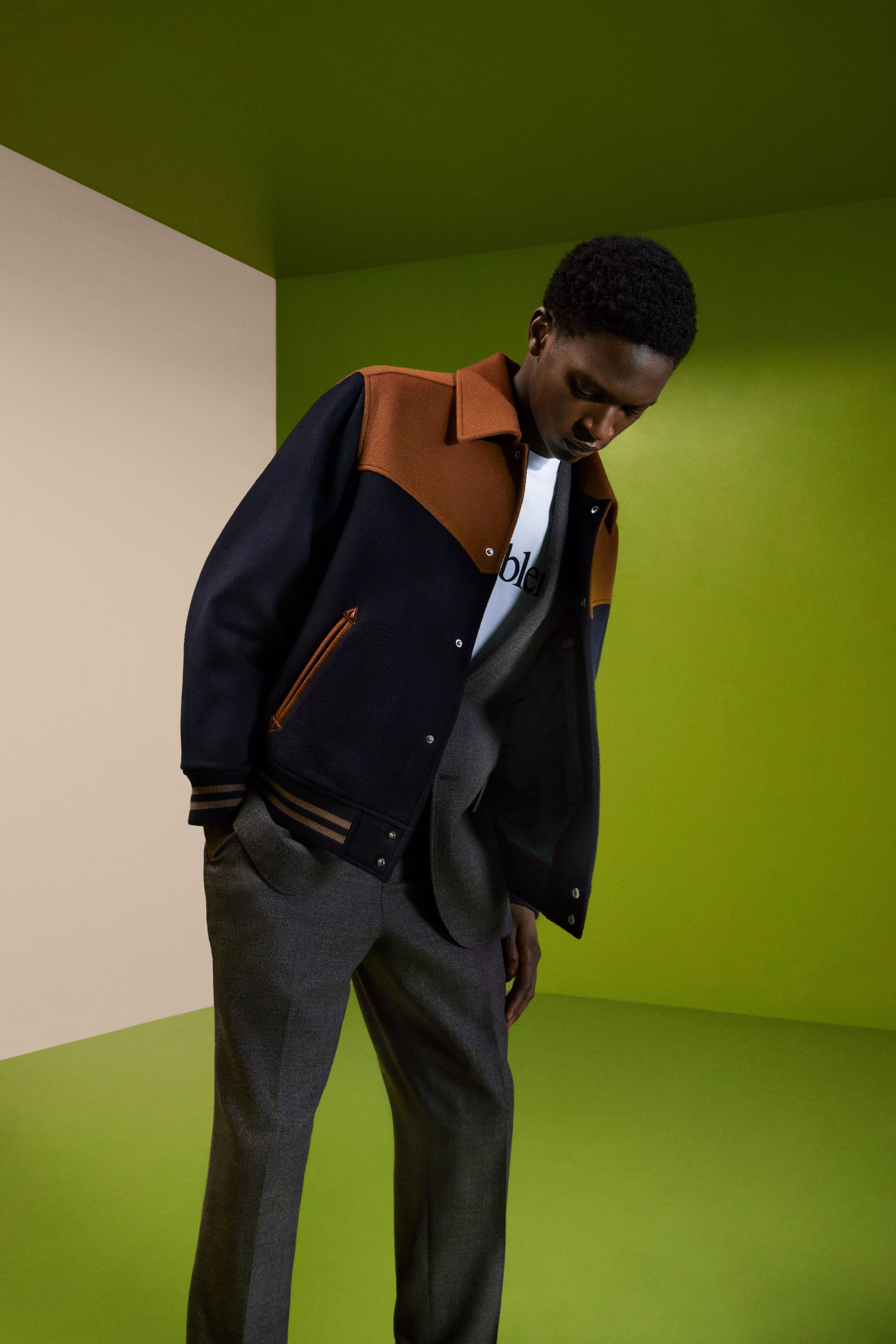 Fashion model wearing a navy and tan varsity jacket with grey trousers in a modern green studio setting