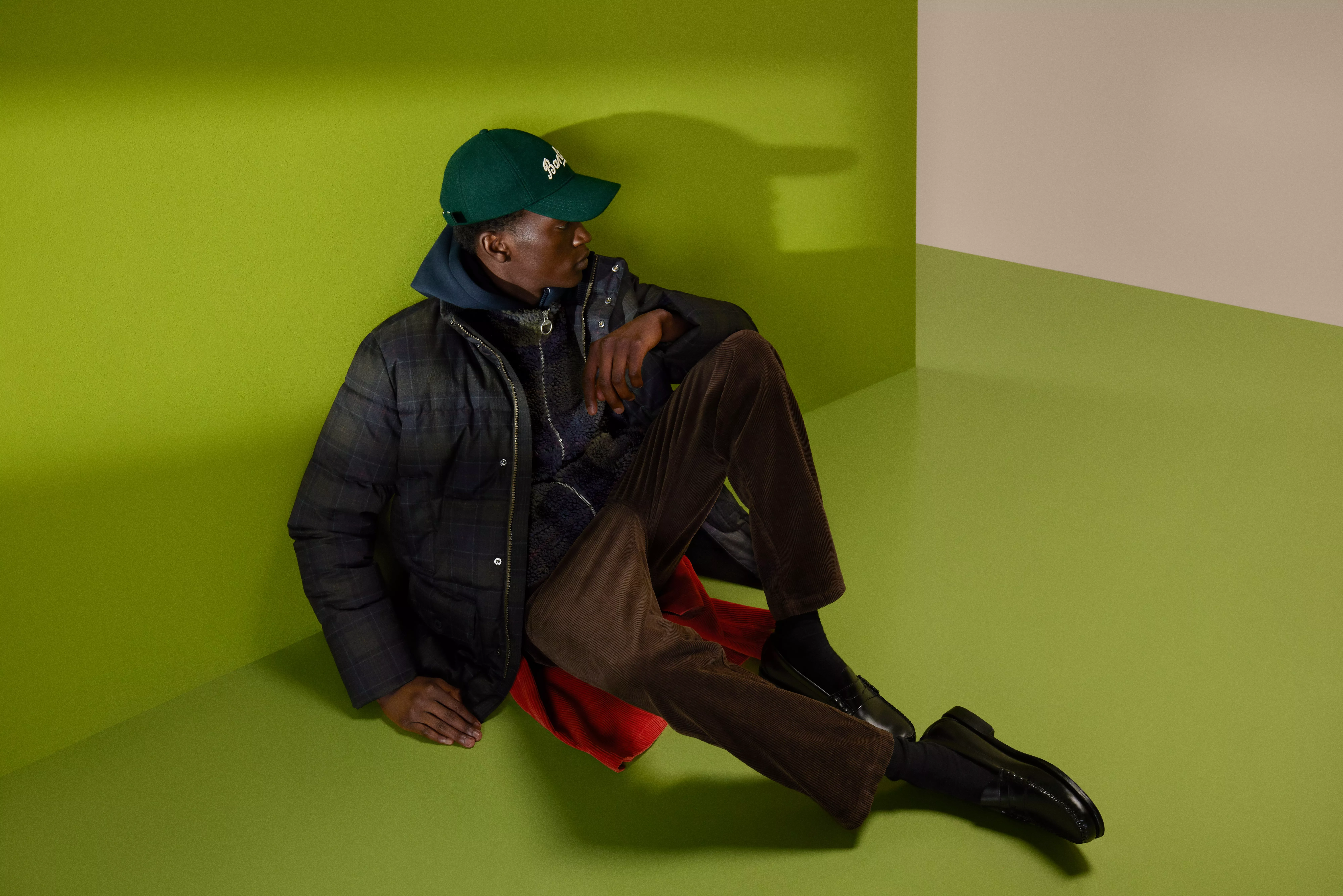 Fashion portrait of man in cap and quilted jacket, sitting on green studio set, wearing cord trousers and loafers
