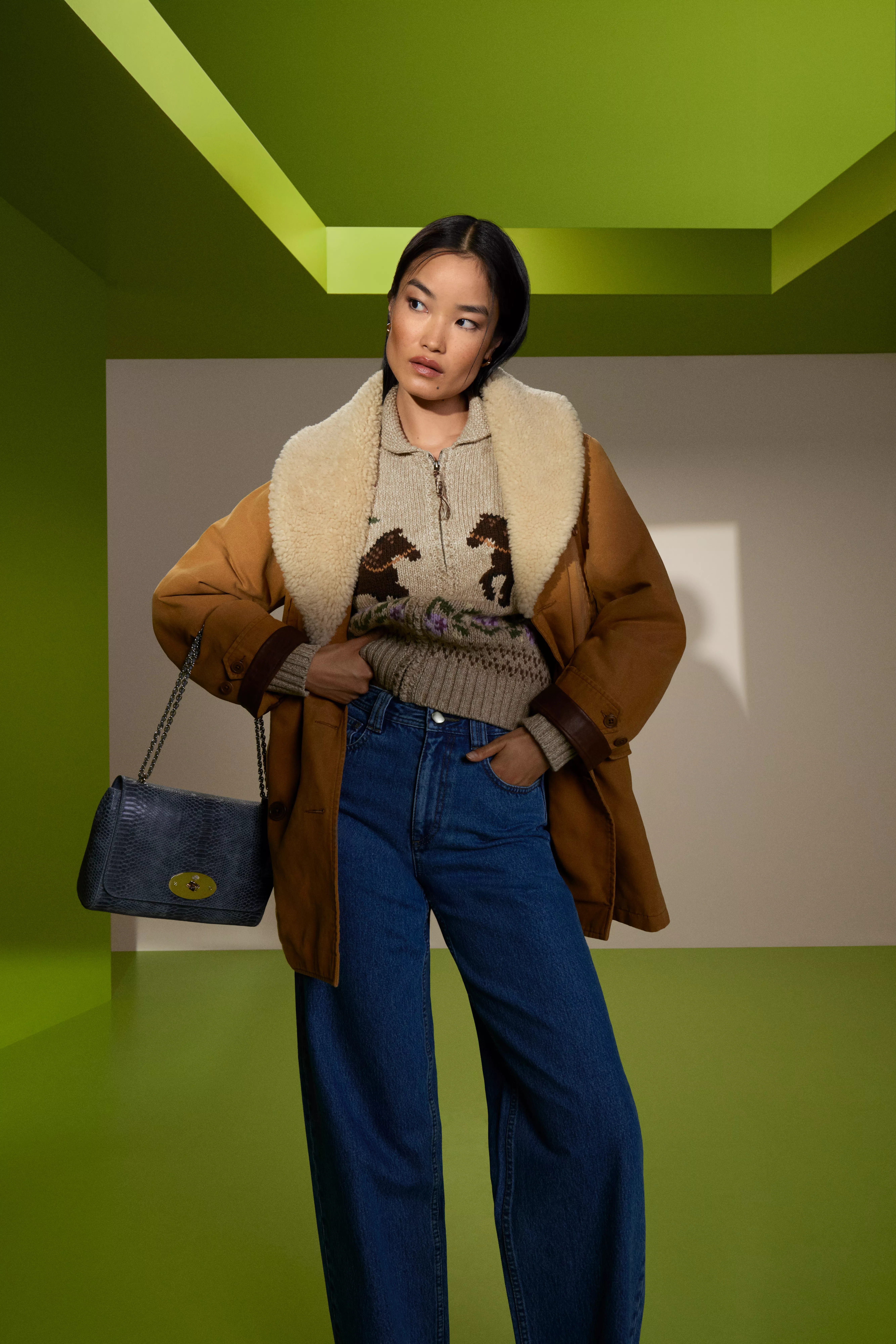 Fashion model in shearling-lined tan jacket, patterned knit jumper and wide-leg jeans, holding handbag in studio set