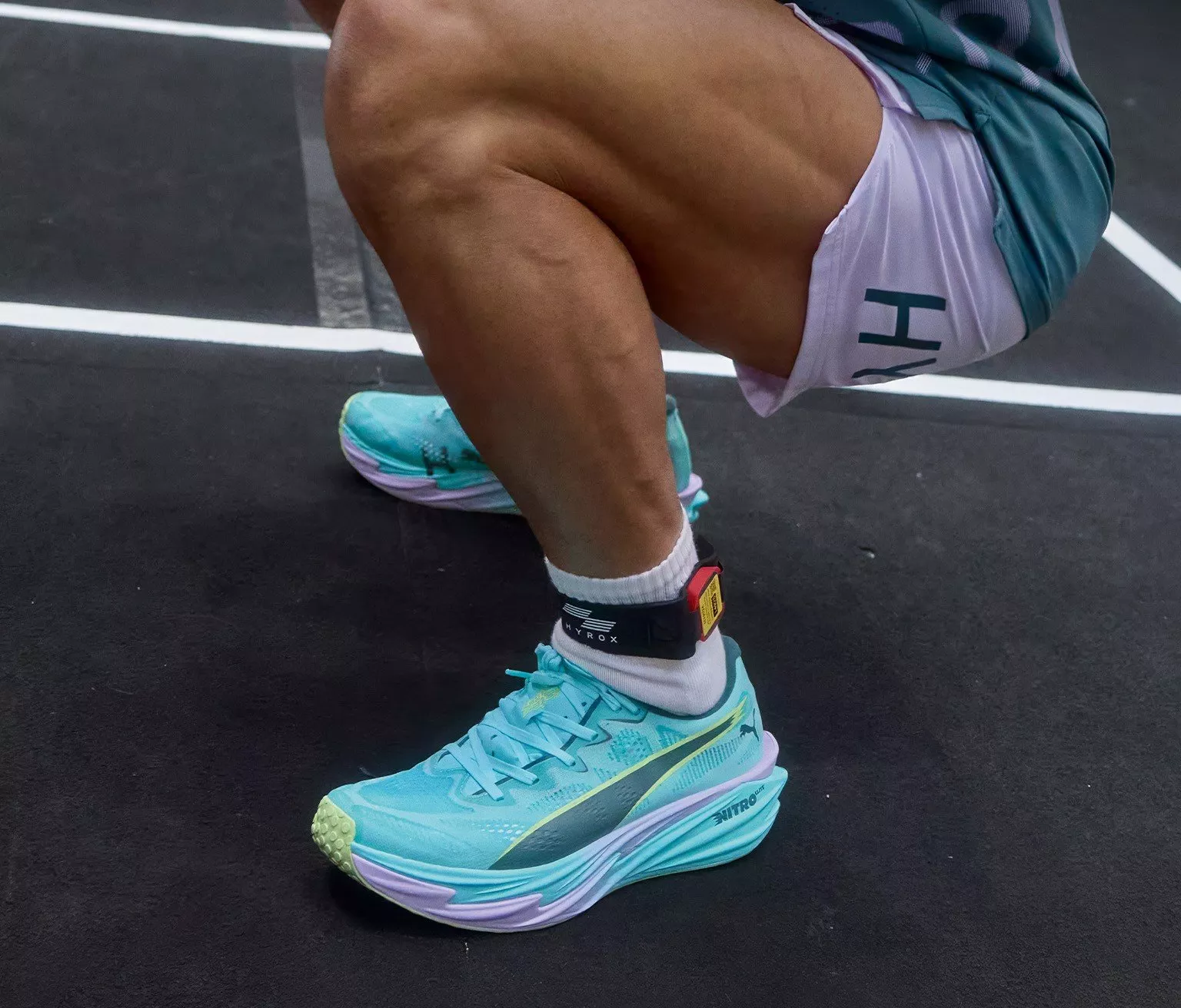 Close-up of athlete squatting on track in turquoise running trainers, socks and ankle sensor strap.