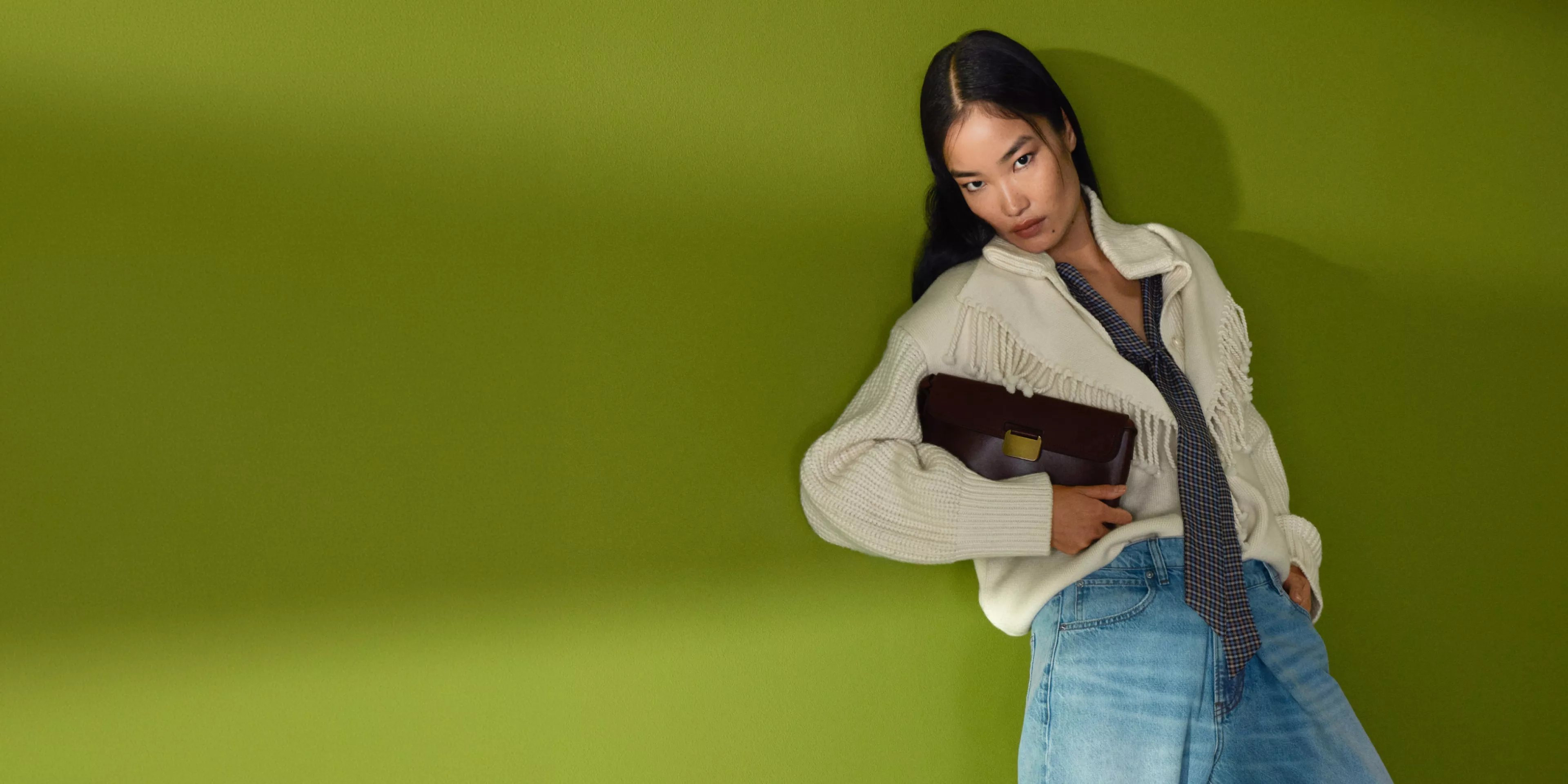 Fashion model in cream fringed knit jumper and denim skirt, holding brown clutch bag against green backdrop