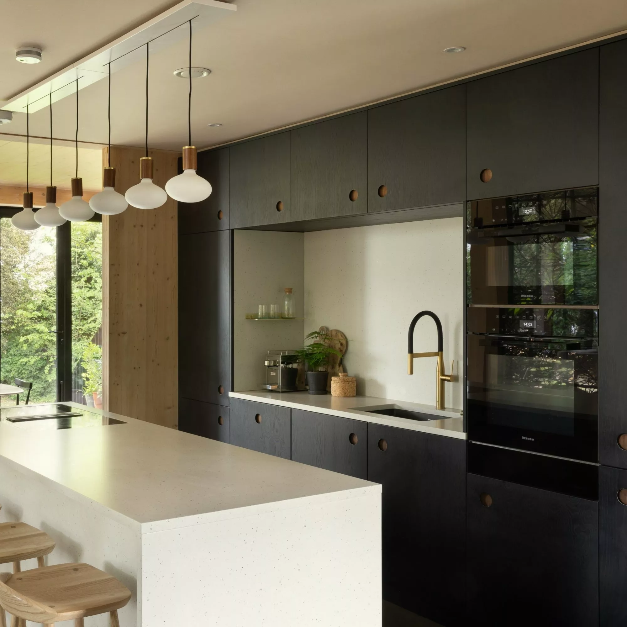 Modern minimalist kitchen with black cabinets, white island, brass tap, pendant lights and garden views through large doors
