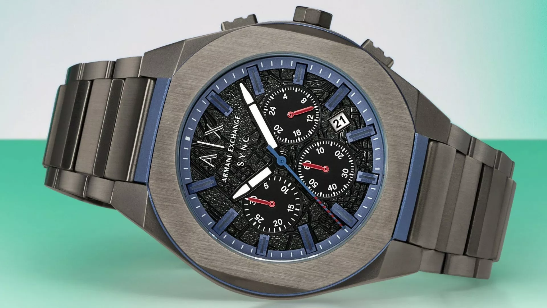 Armani Exchange gunmetal chronograph watch with blue dial accents, link bracelet, and exposed sub-dials set against a mint green background.