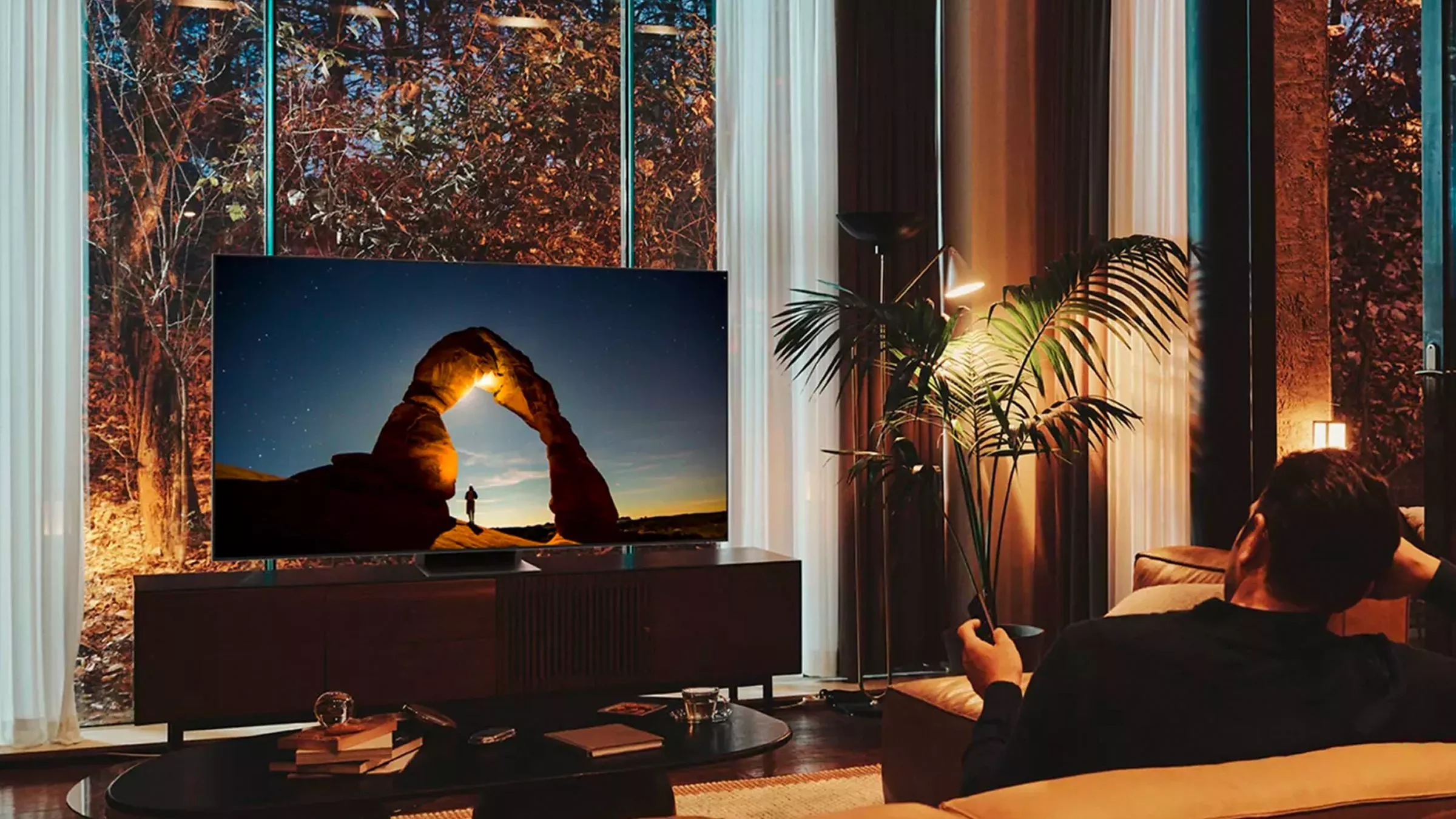 A person relaxes on a sofa, watching a vivid image of a sunset arch on a large television in a stylish living room.