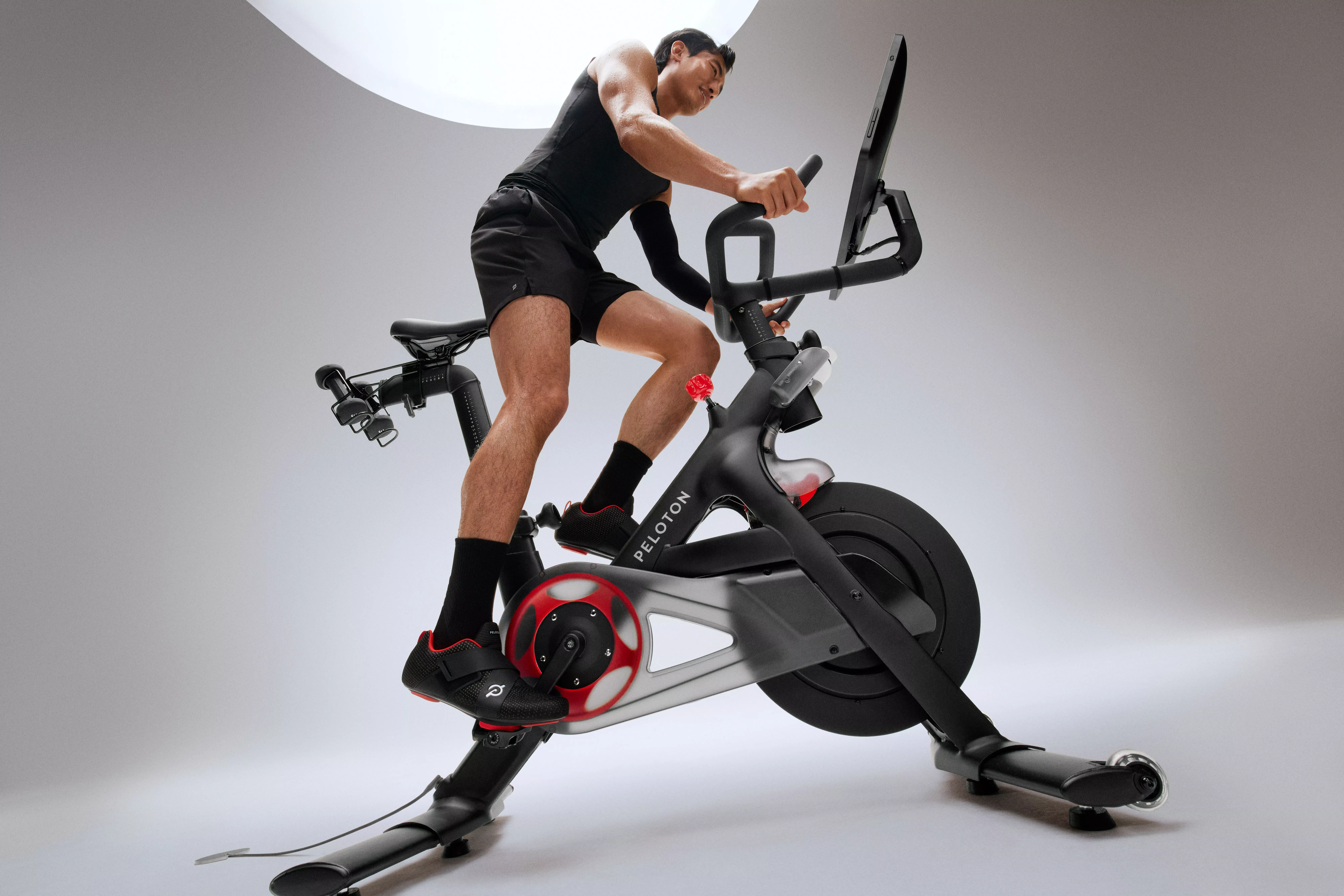 Man using Peloton exercise bike in studio, indoor cycling workout with touchscreen and resistance flywheel.