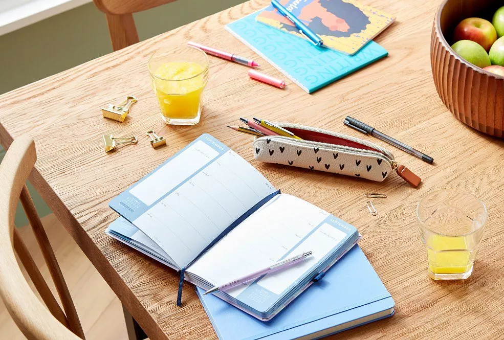 Open weekly planner and pencil case on wooden table with pens, notebooks, paper clips and orange juice glasses