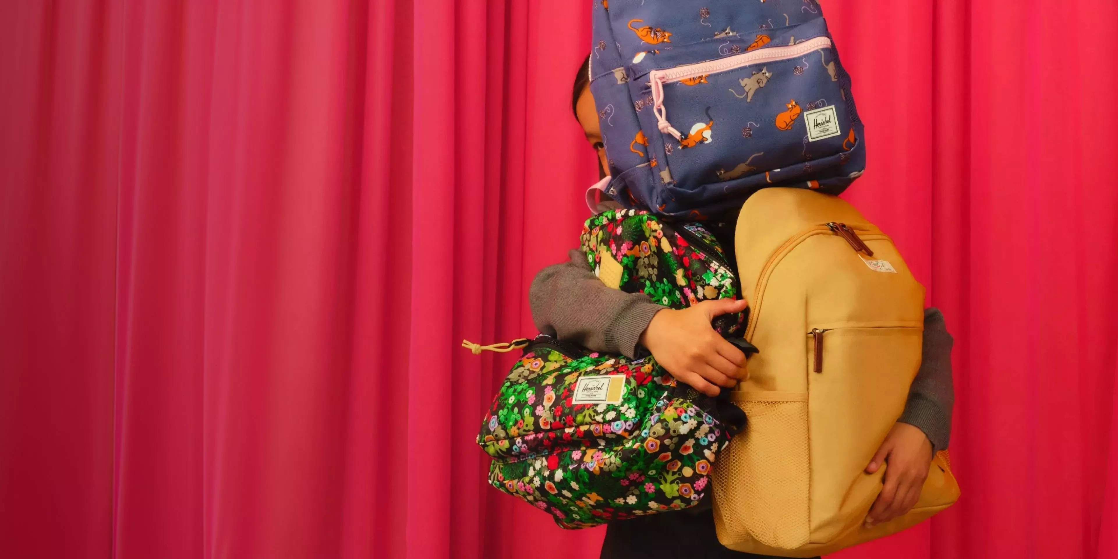 kid carrying loads of backpacks with a red curtain behind her