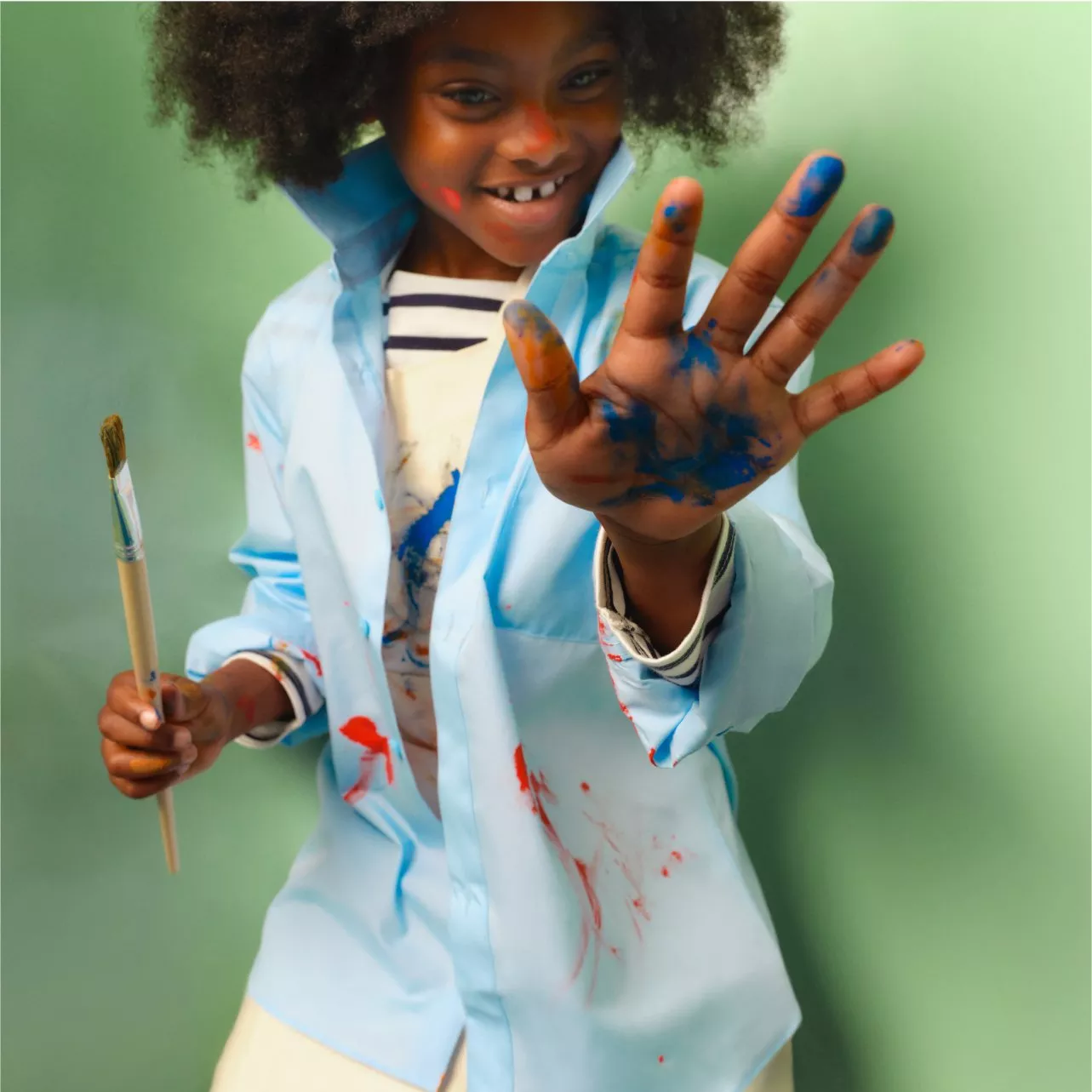 boy with paint on his hand and face, holding a paint brush and wearing a light blue shirt with paint stains