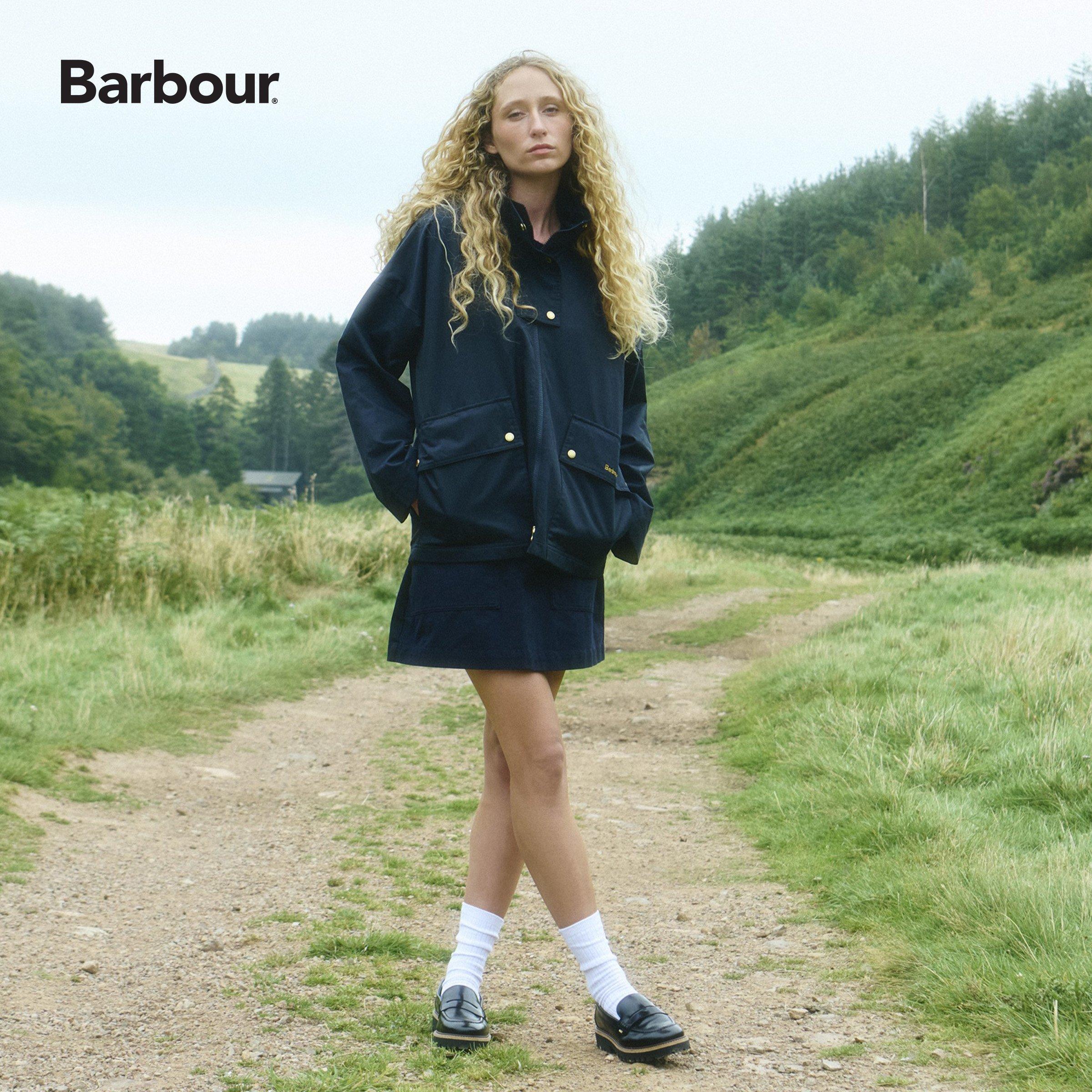 A woman with curly blonde hair standing on a dirt path in a green, hilly landscape, wearing a navy jacket, skirt, white socks, and black shoes.