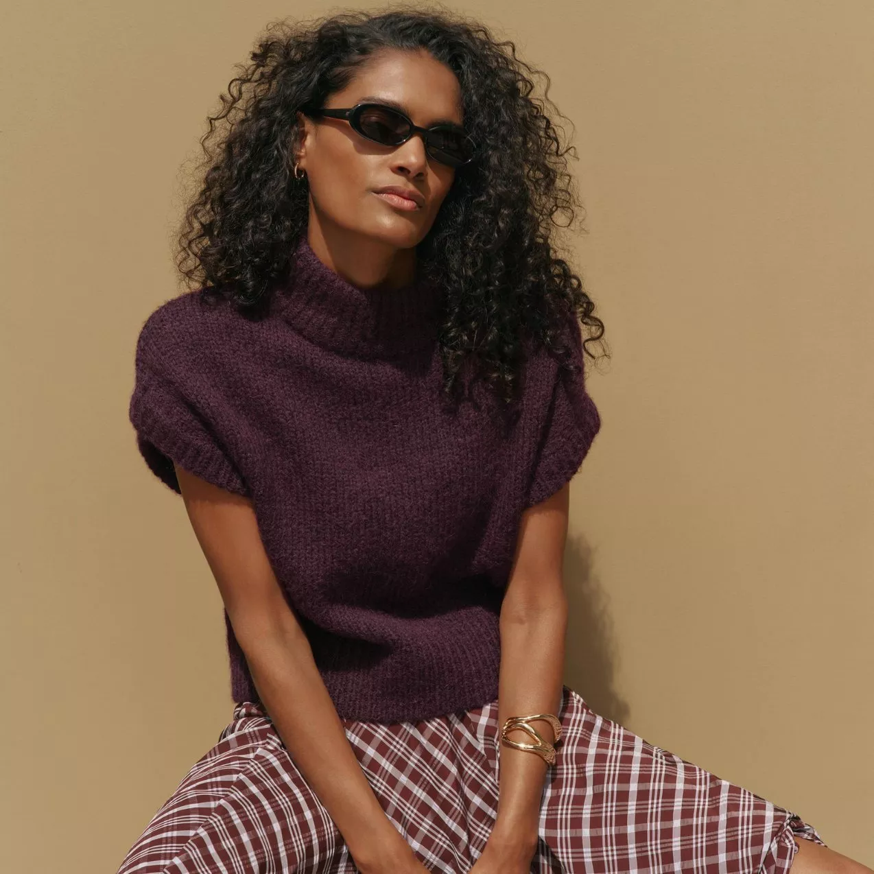 A woman poses wearing black sunglasses, a cap-sleeved dark purple high-neck knit sweater, a gold bracelet, and a brown plaid skirt against a beige background.