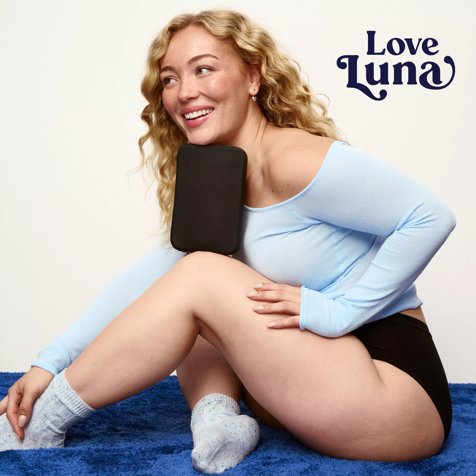Smiling woman in light blue top holding black Love Luna period underwear case, seated on blue rug.