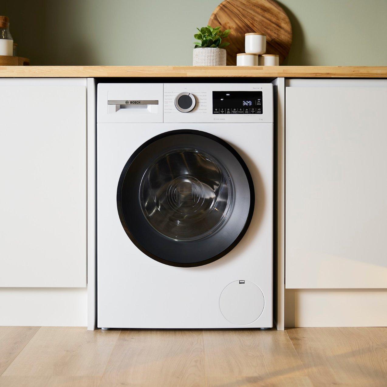 Modern front-loading white washing machine with a black door frame, integrated beneath a light wooden countertop between modern kitchen cabinets.