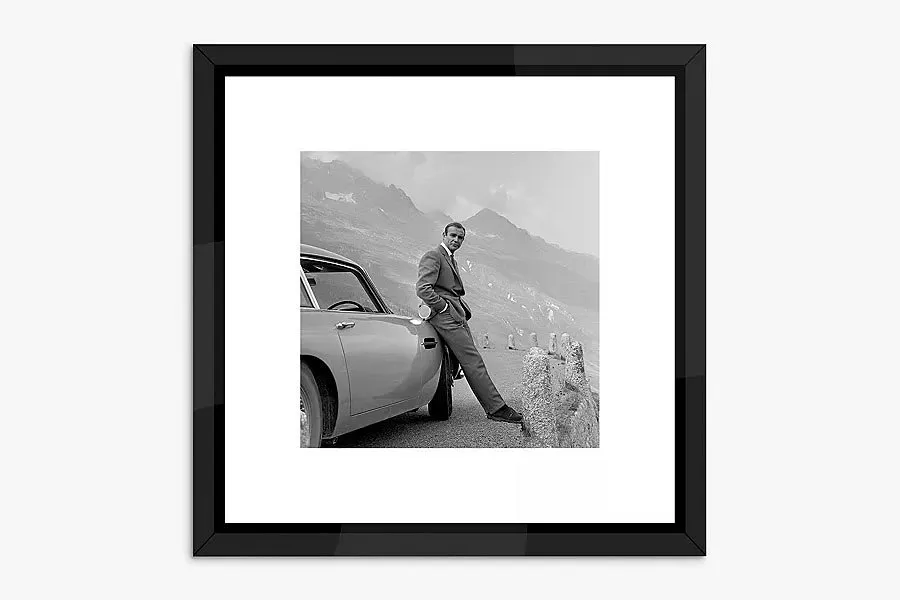 Framed black and white photo of a man leaning on a classic car on a mountain road viewpoint