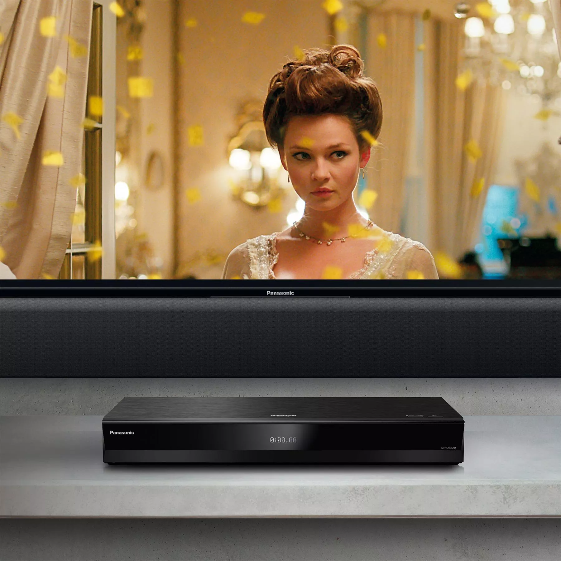 A black Panasonic Blu-ray player sitting on a counter below a TV screen playing a period drama movie with falling gold confetti.